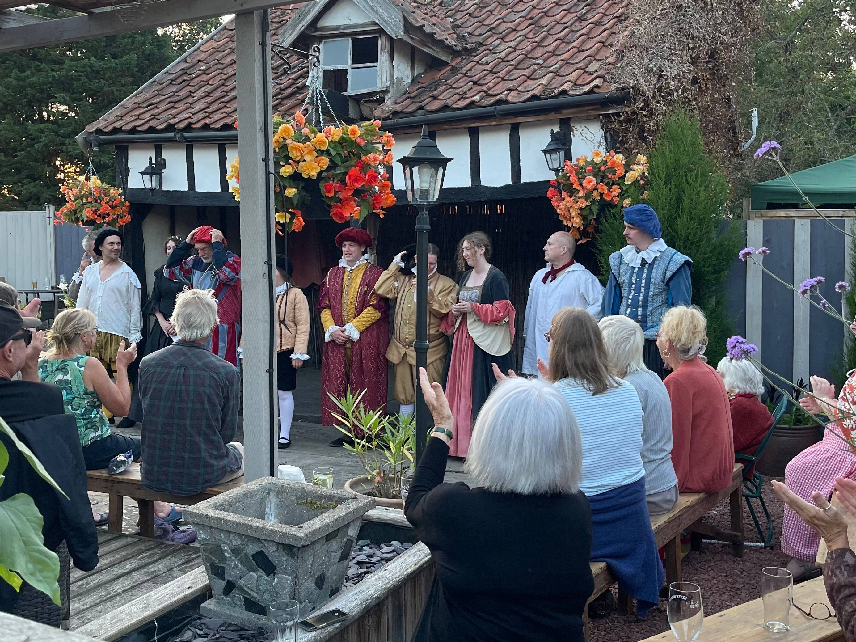Outdoor audience applauding actors in historical costumes on stage outside a rustic building.