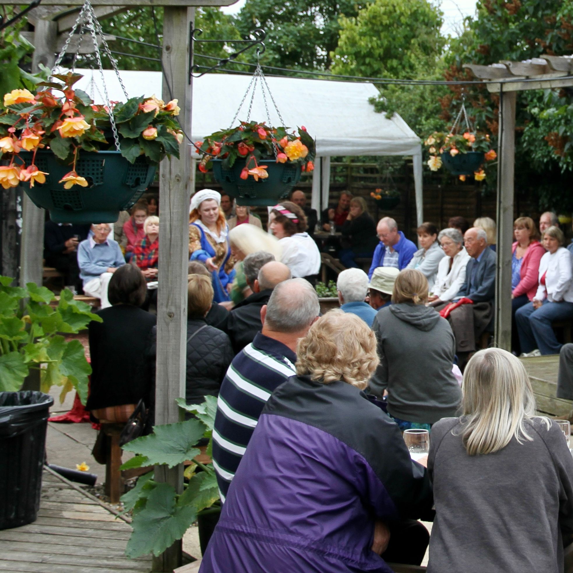 A group of people gathered outdoors under wooden pergolas, with hanging baskets of flowers, watching a performance where some people are dressed in historical costumes.