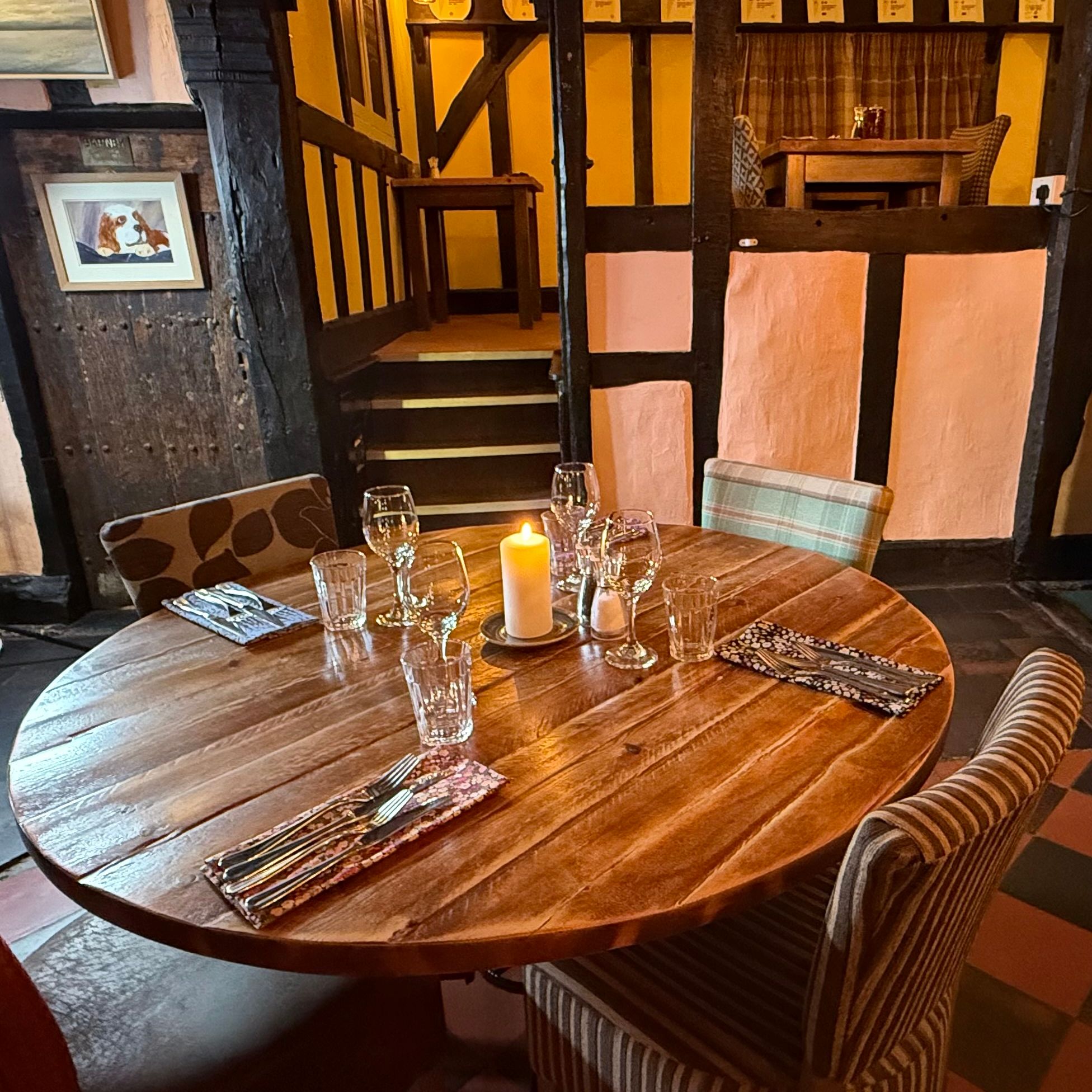 Cozy rustic dining area with a round wooden table set for four, surrounded by upholstered chairs in a warm, timber-framed room.