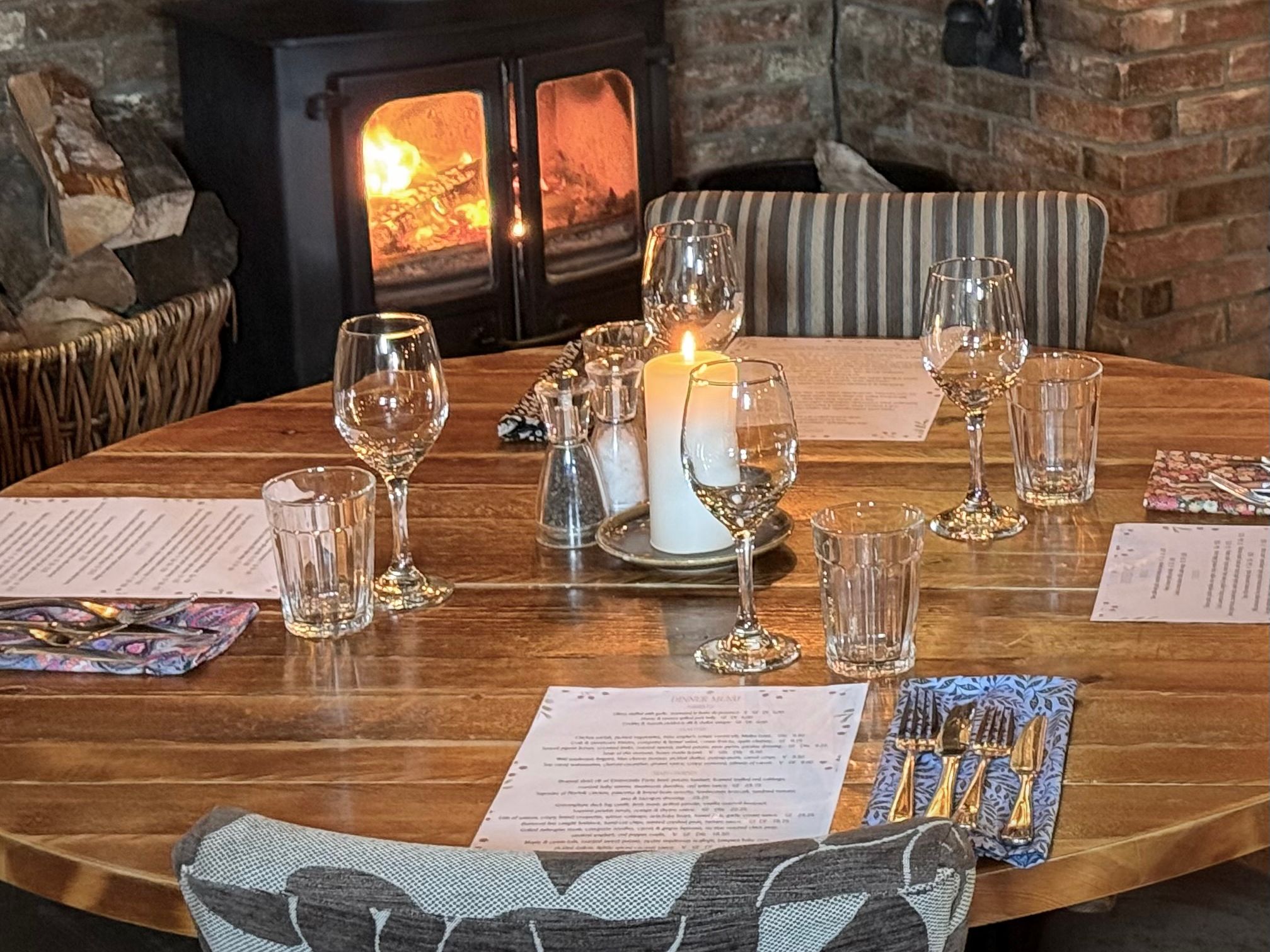 Cozy restaurant table set with glassware and a lit candle in front of a wood-burning stove and brick fireplace