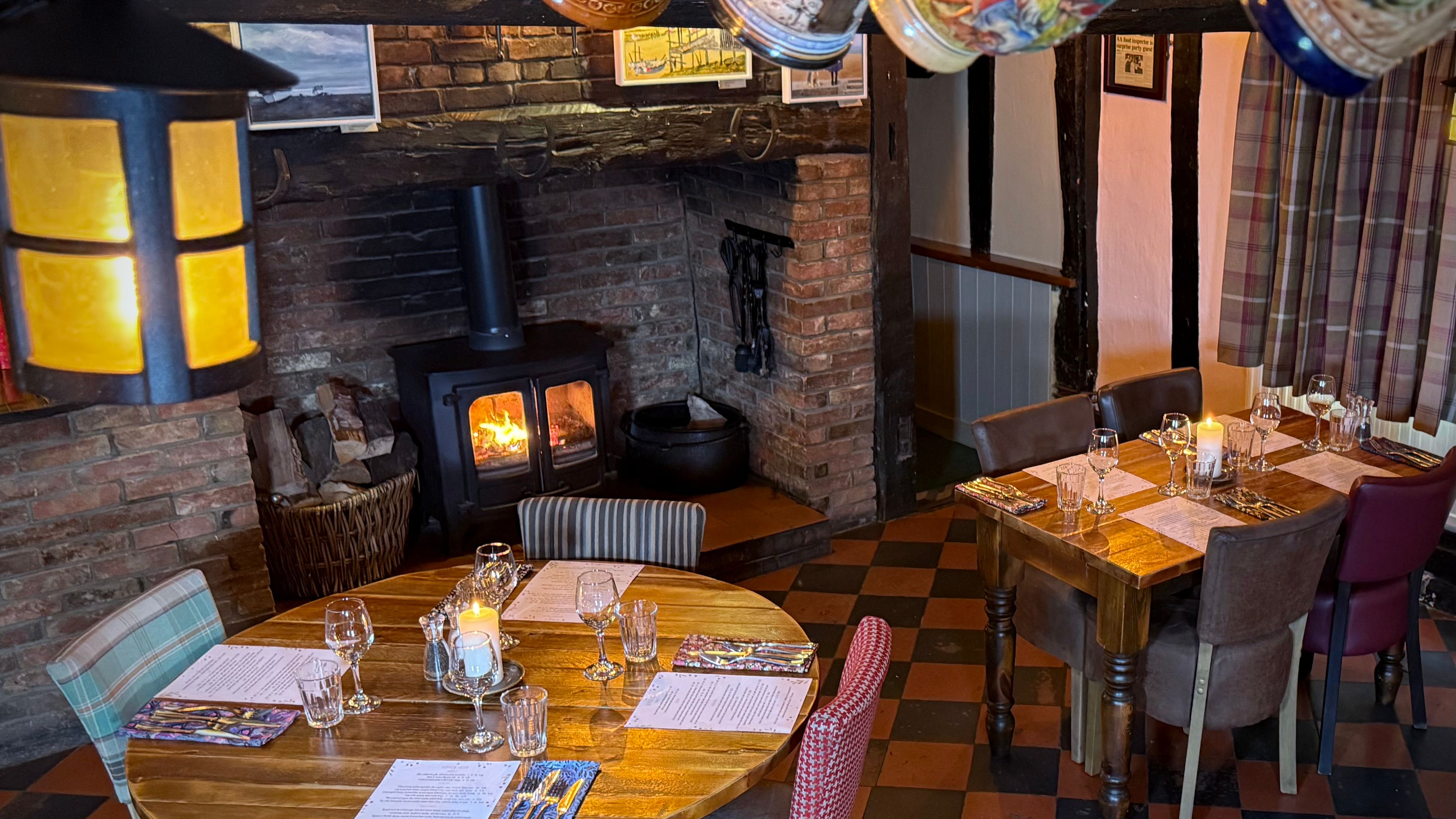 Cozy restaurant interior with a fireplace, wooden tables set for dining, and decorative mugs hanging from a ceiling beam.