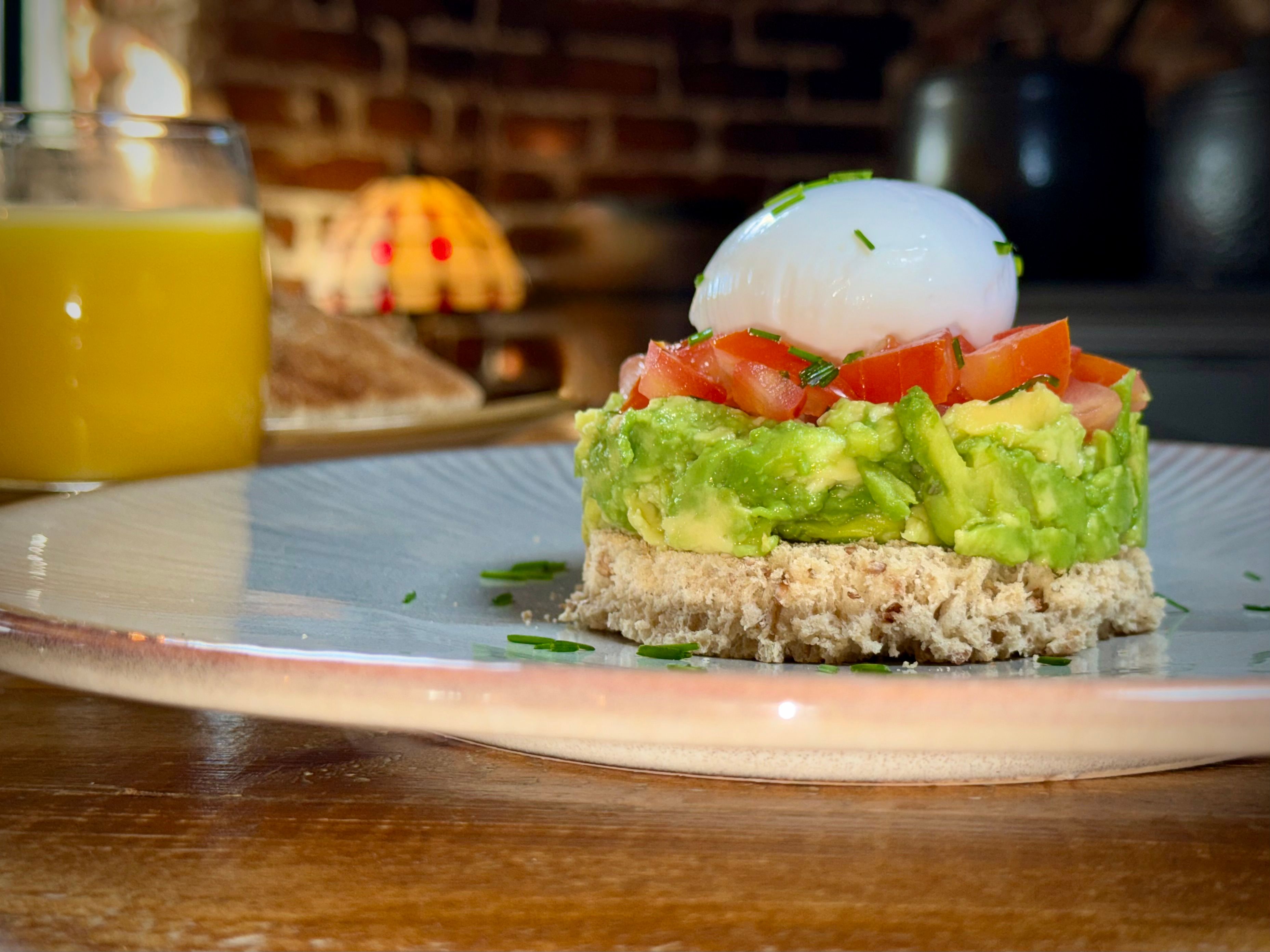 A breakfast plate with a stack of bread, mashed avocado, diced tomatoes, and a poached egg, with a glass of orange juice in the background.