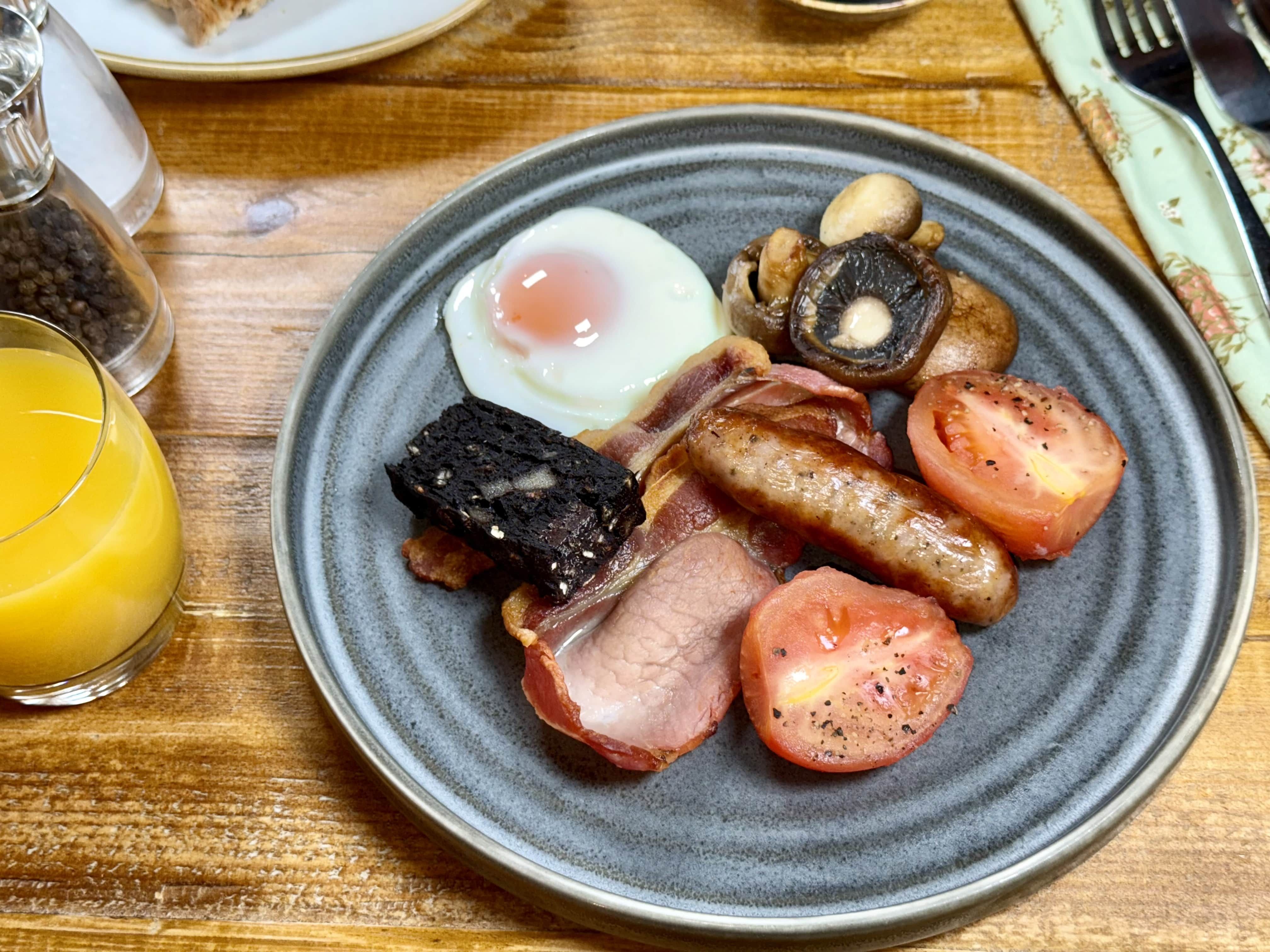 Traditional English breakfast with bacon, sausage, egg, black pudding, mushrooms, and tomatoes on a grey plate with a glass of orange juice.