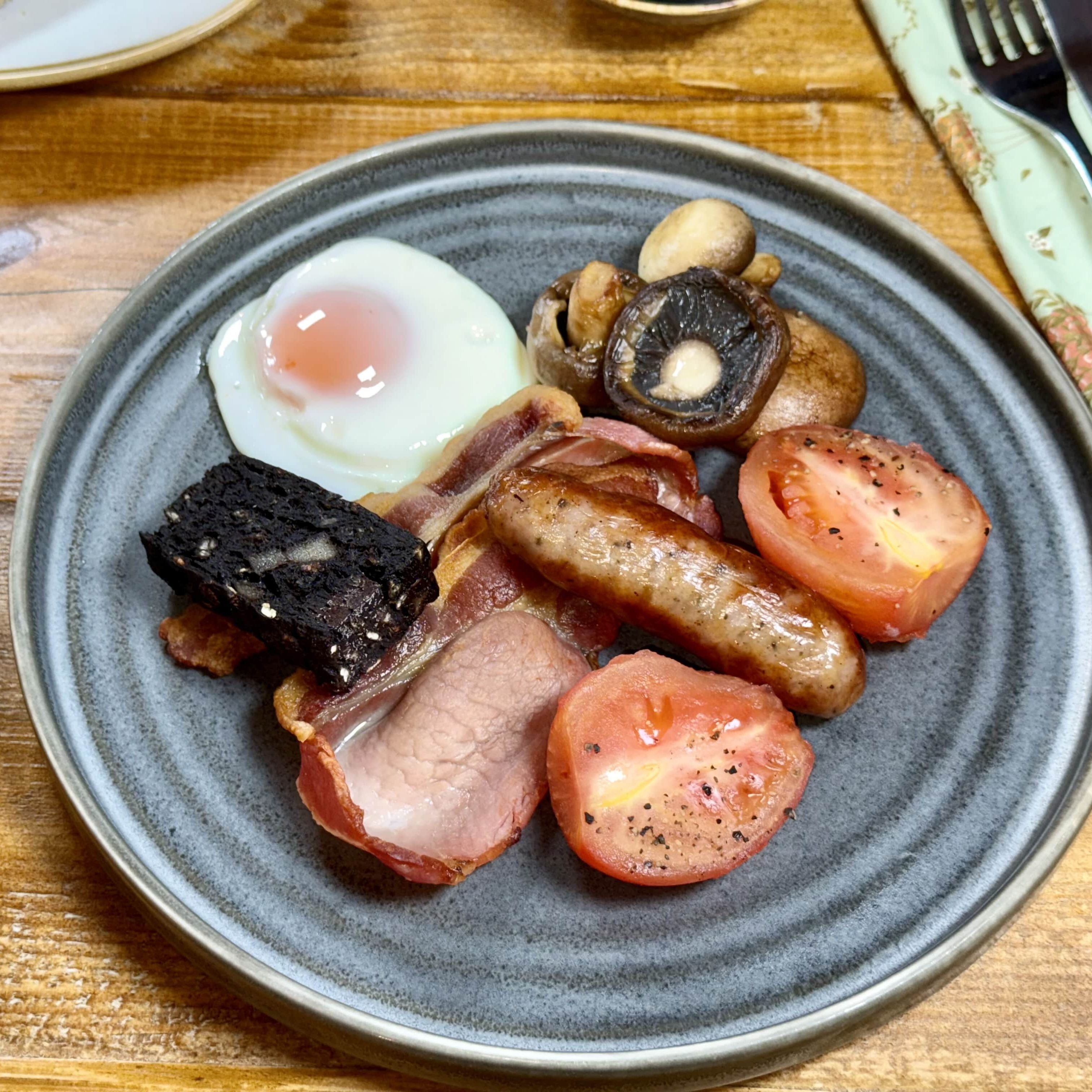 Traditional English breakfast with bacon, sausage, egg, black pudding, mushrooms, and tomatoes on a grey plate with a glass of orange juice.