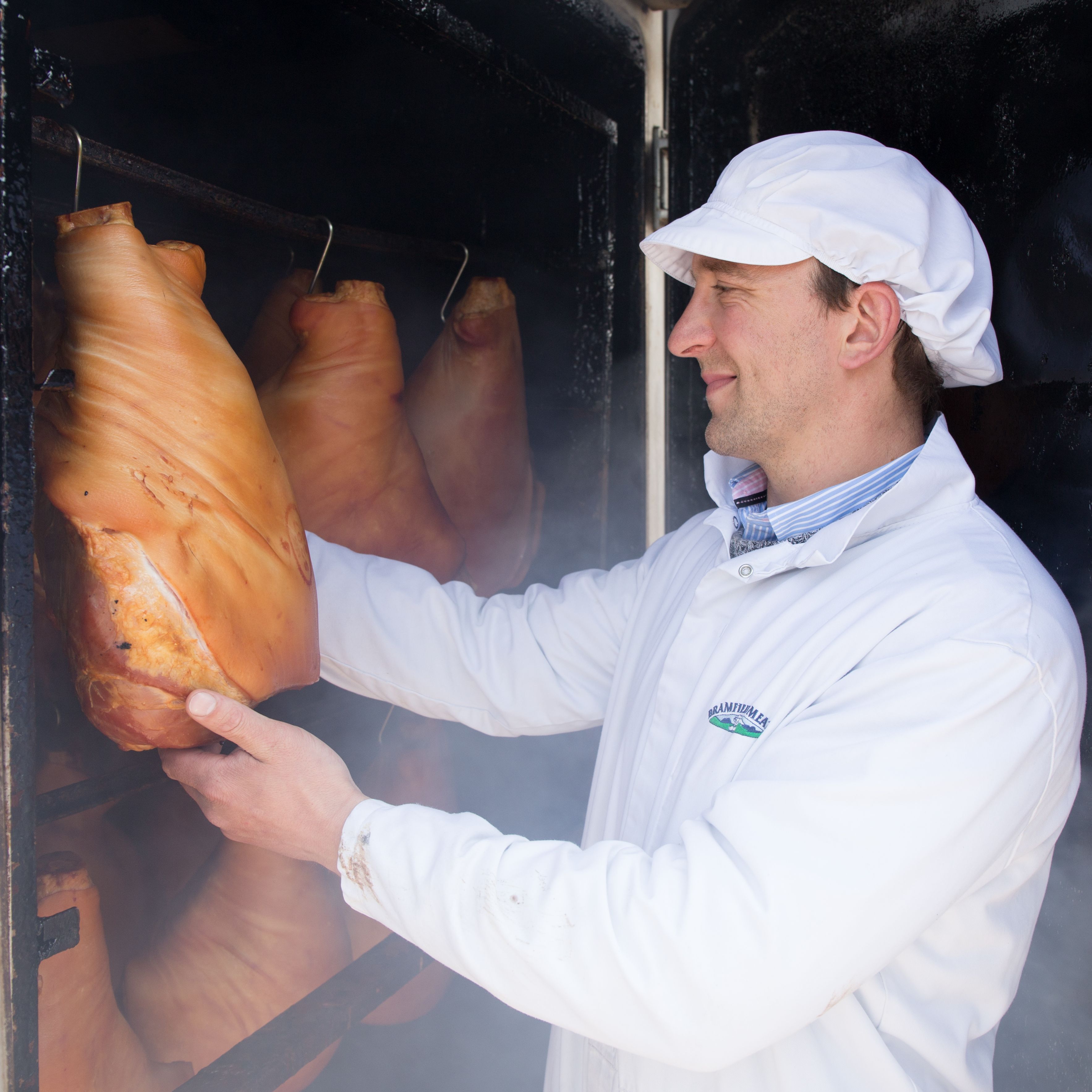 Man in white coat and cap inspecting smoked meat hanging in a smokehouse