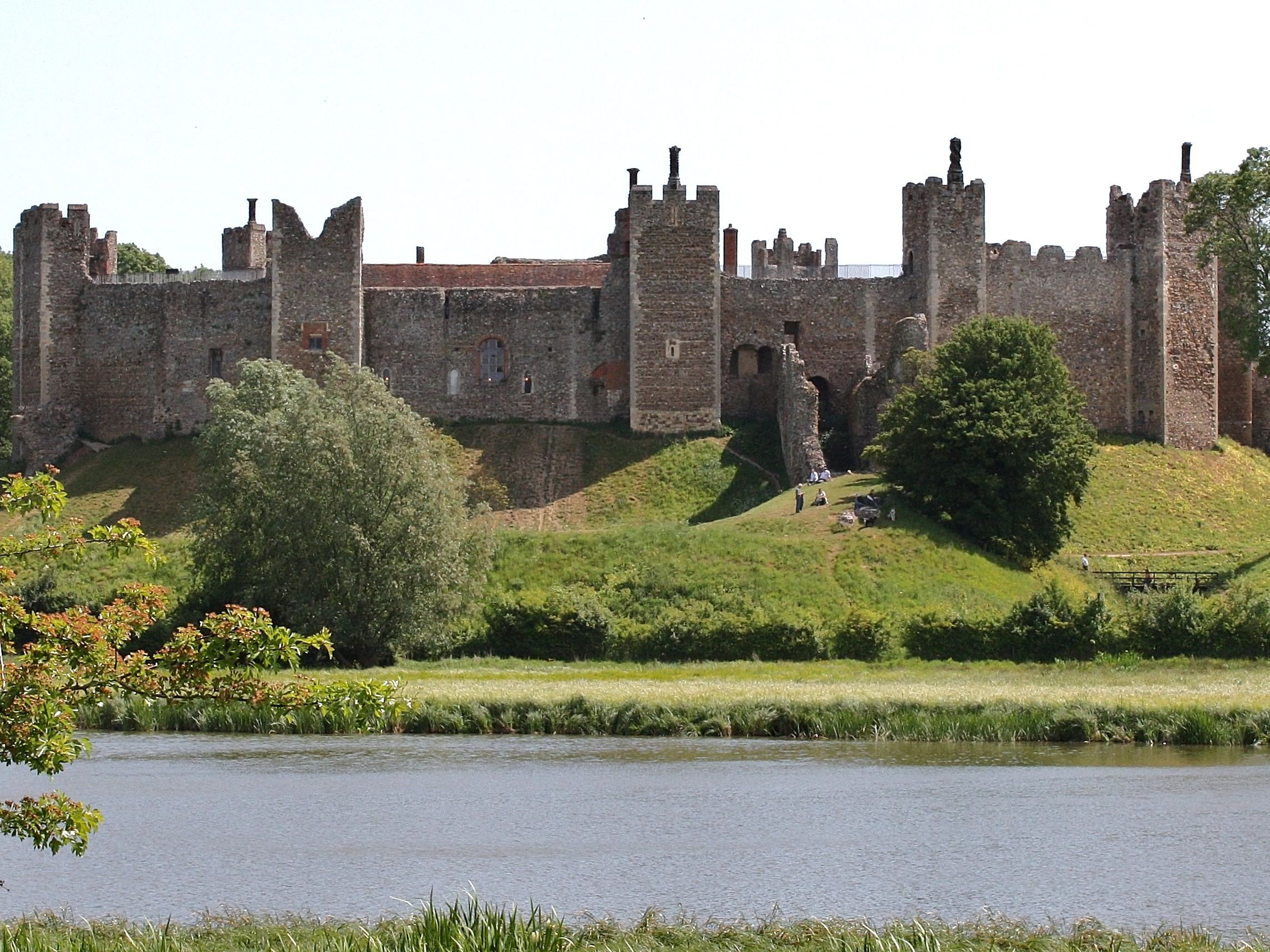 A large historic stone castle with multiple towers and walls, situated on a grassy hill beside a river.