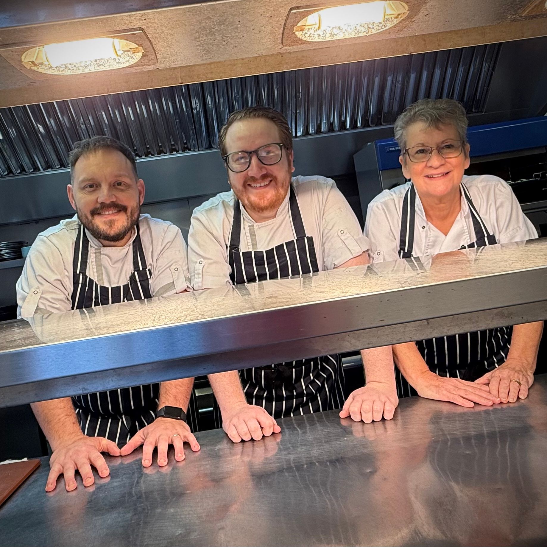 Three chefs in white uniforms and black-striped aprons smiling behind a counter in a commercial kitchen.