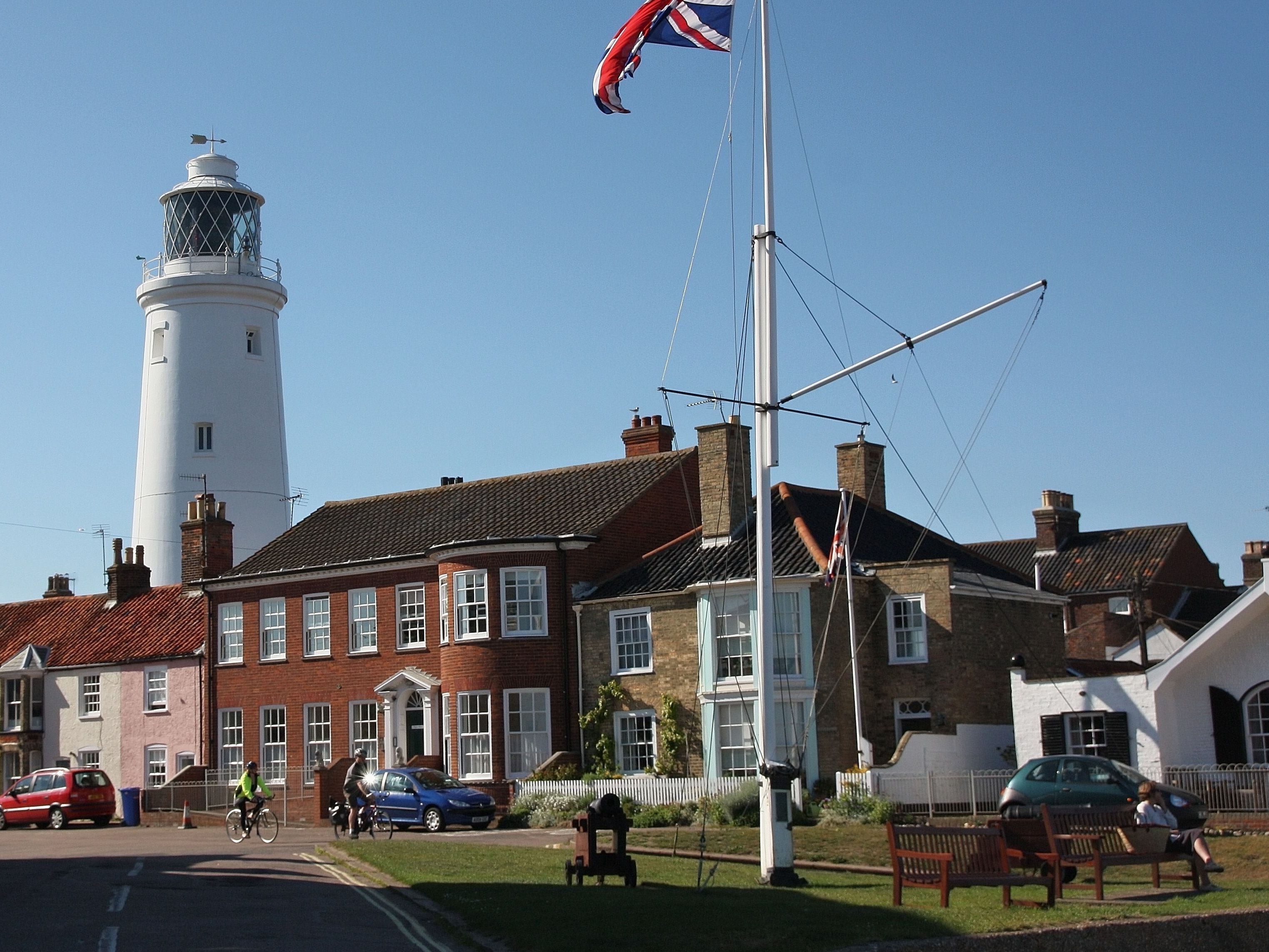 A white lighthouse behind residential houses, with a flagpole flying the Union Jack and people sitting on benches and riding bicycles.