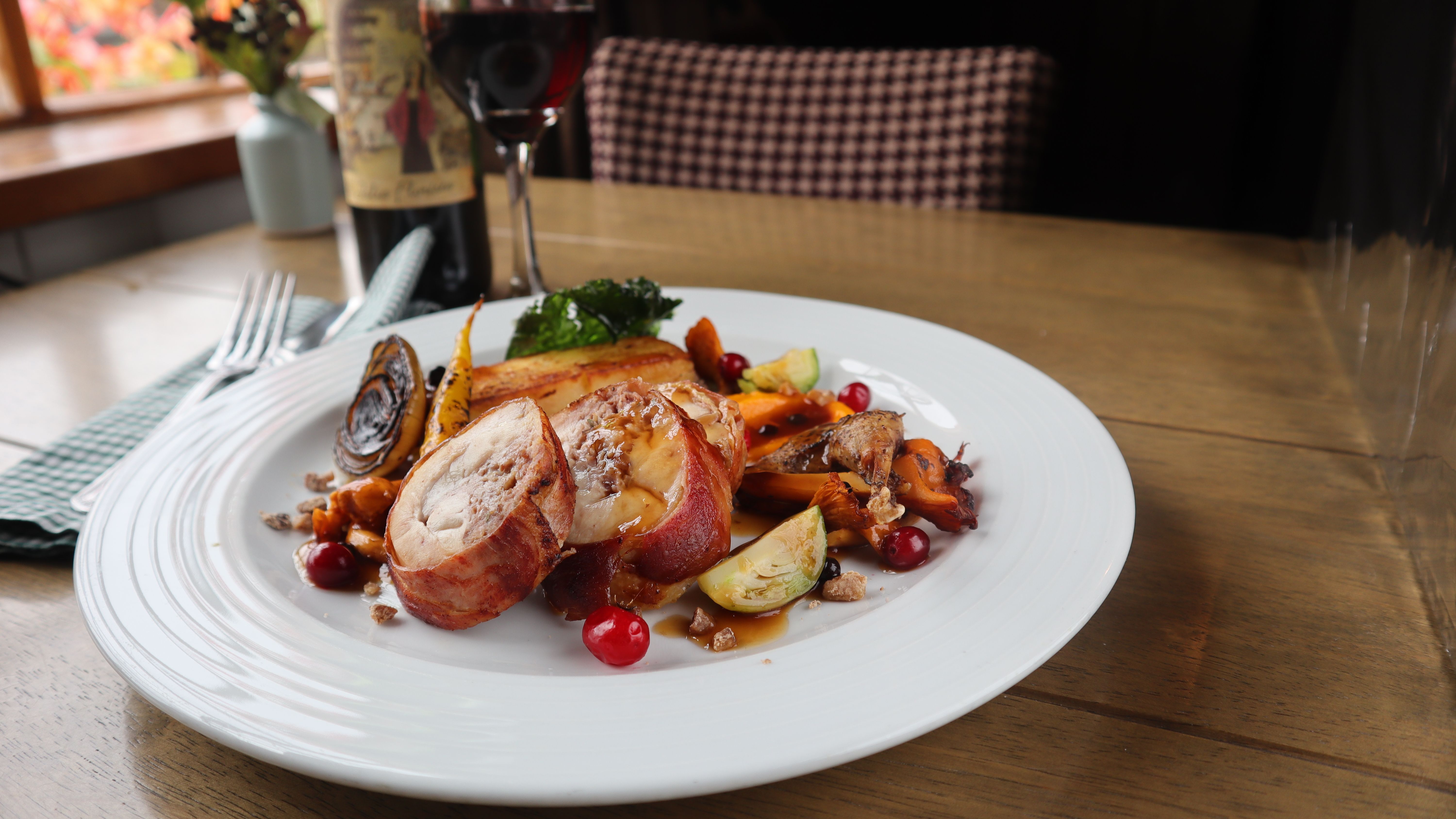Gourmet dinner plate with rolled meat, roasted vegetables, and berries, with a glass of red wine in the background.