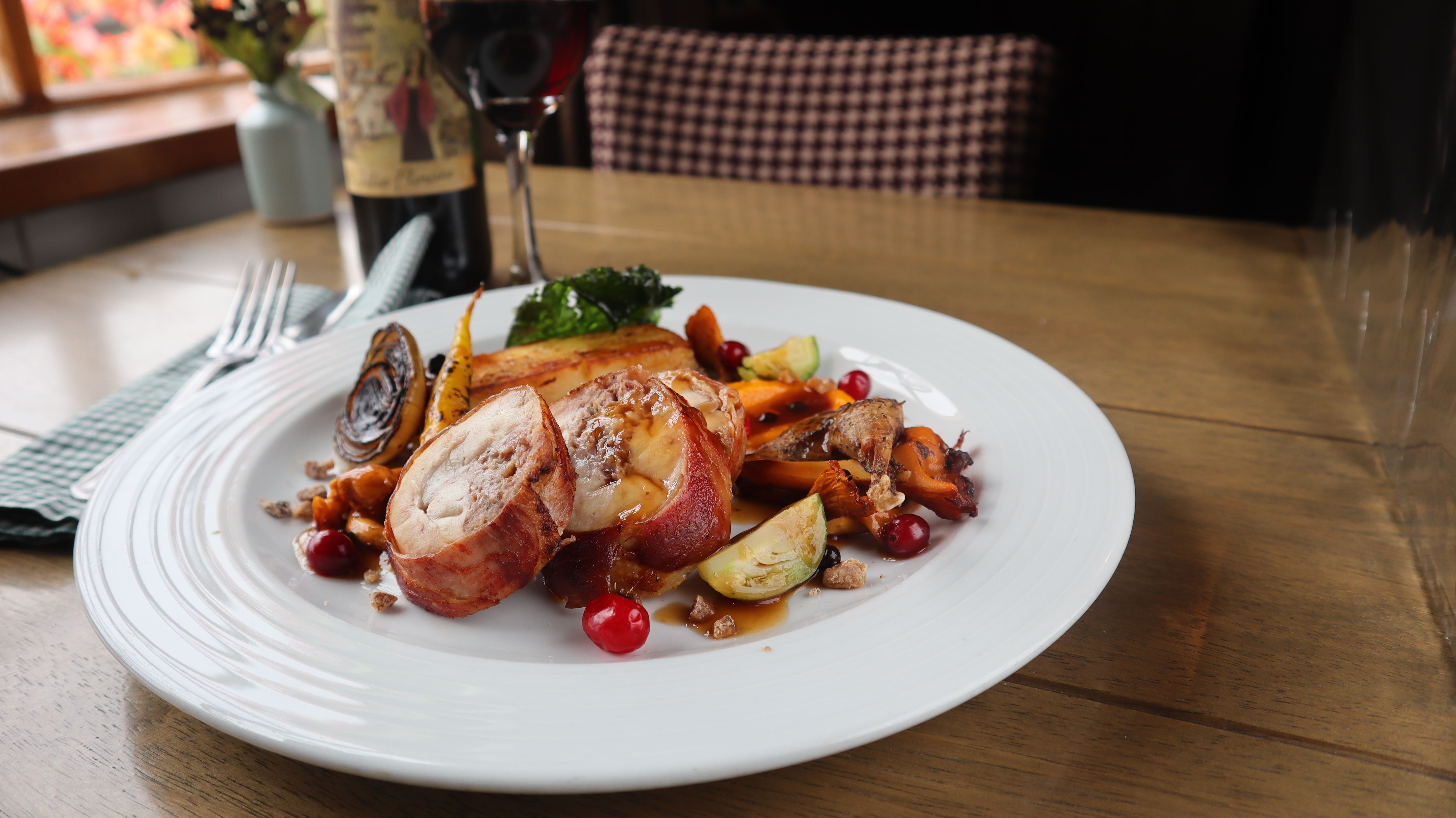 Gourmet dinner plate with rolled meat, roasted vegetables, and berries, with a glass of red wine in the background.