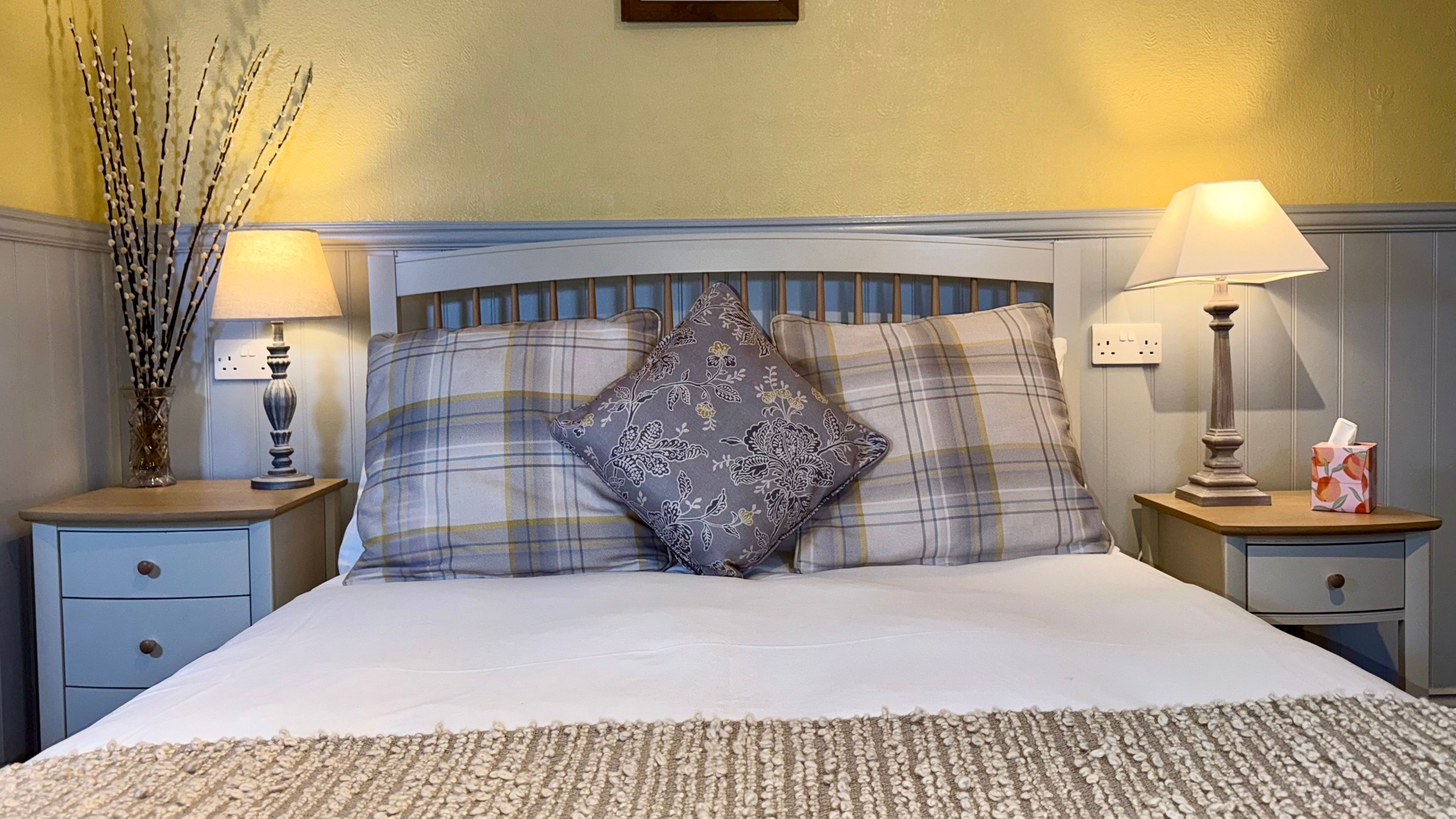 Cozy bedroom with double bed, plaid and decorative pillows, bedside tables with lamps, and wall art above the bed.