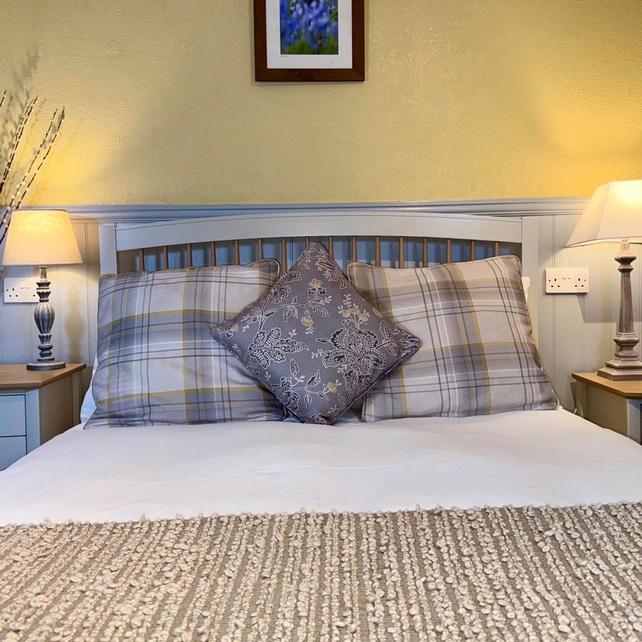 Cozy bedroom with double bed, plaid and decorative pillows, bedside tables with lamps, and wall art above the bed.