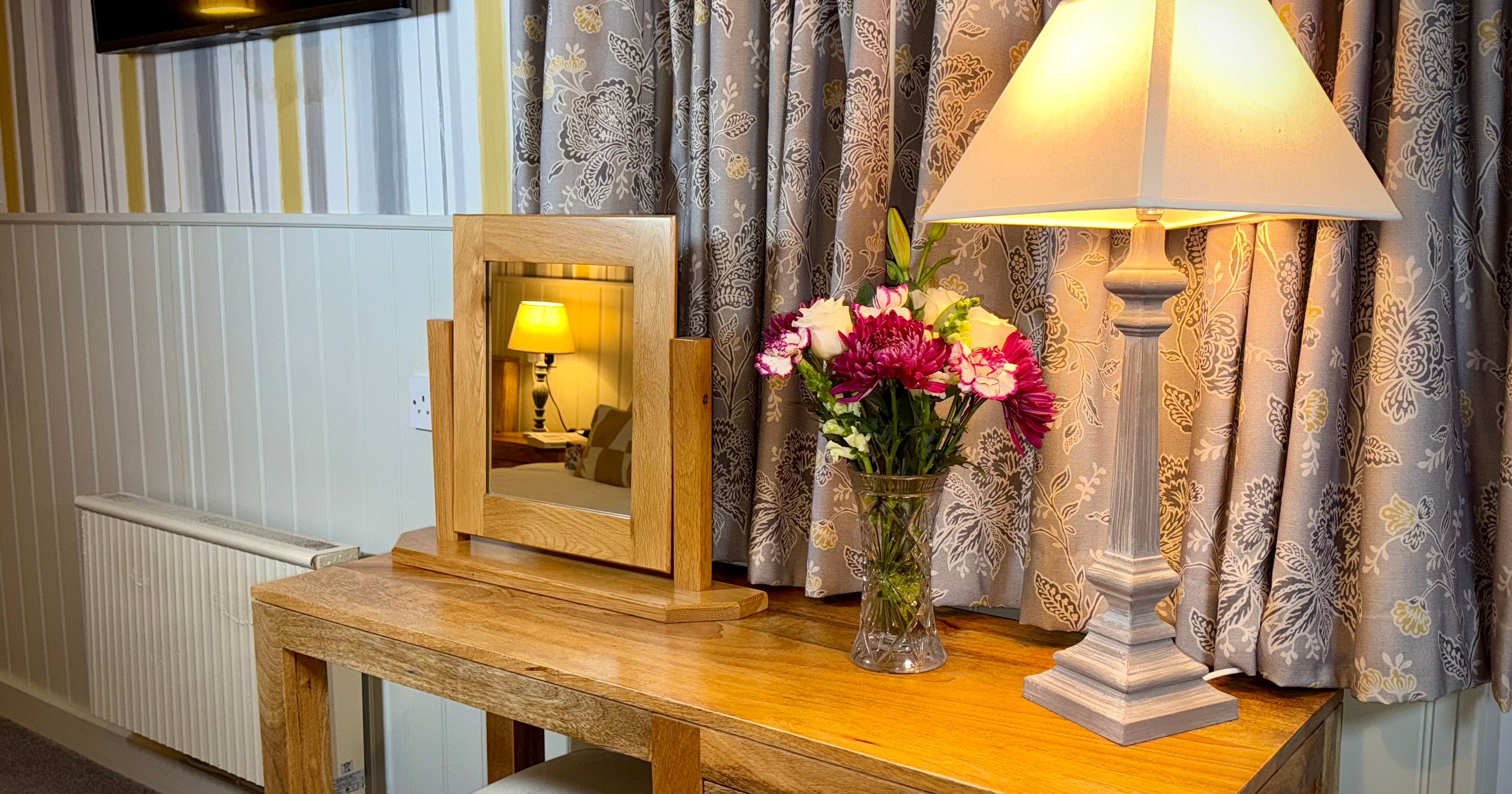Wooden dressing table with a mirror, lamp, and vase of flowers in a cozy room.