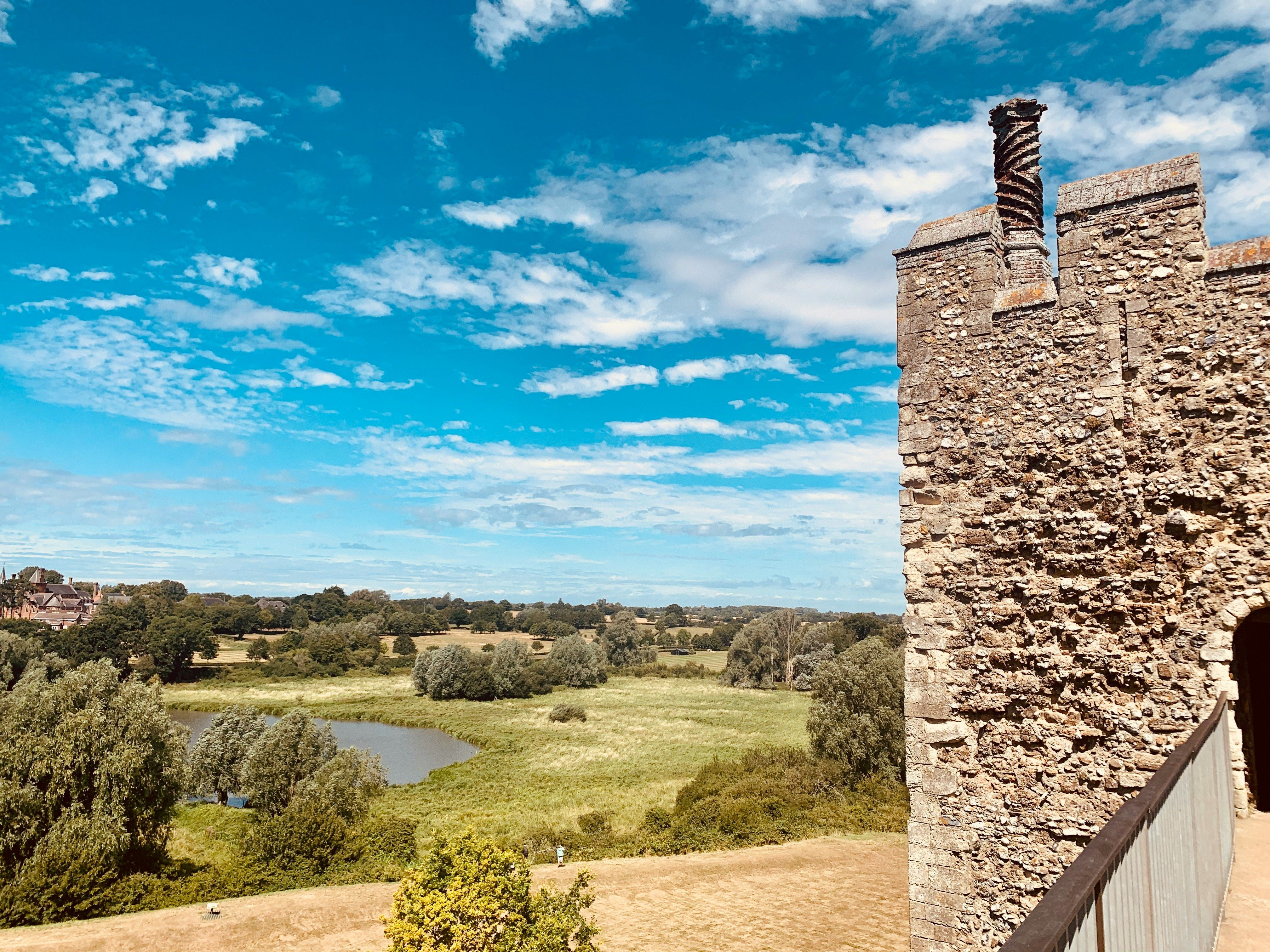Stone wall and battlements of Framlingham Castle overlooking green fields and a pond under a blue sky with scattered clouds.