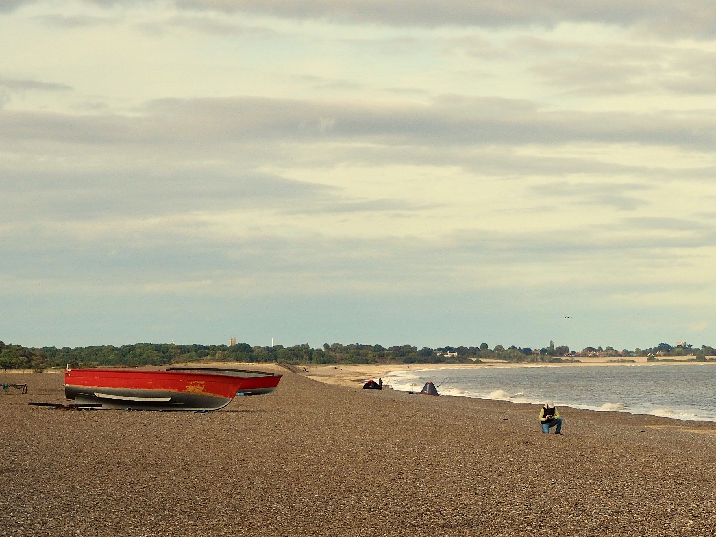 A pebble beach with some fishing boats on the shore, a few people sitting and walking, and the sea stretching to the horizon under a cloudy sky.