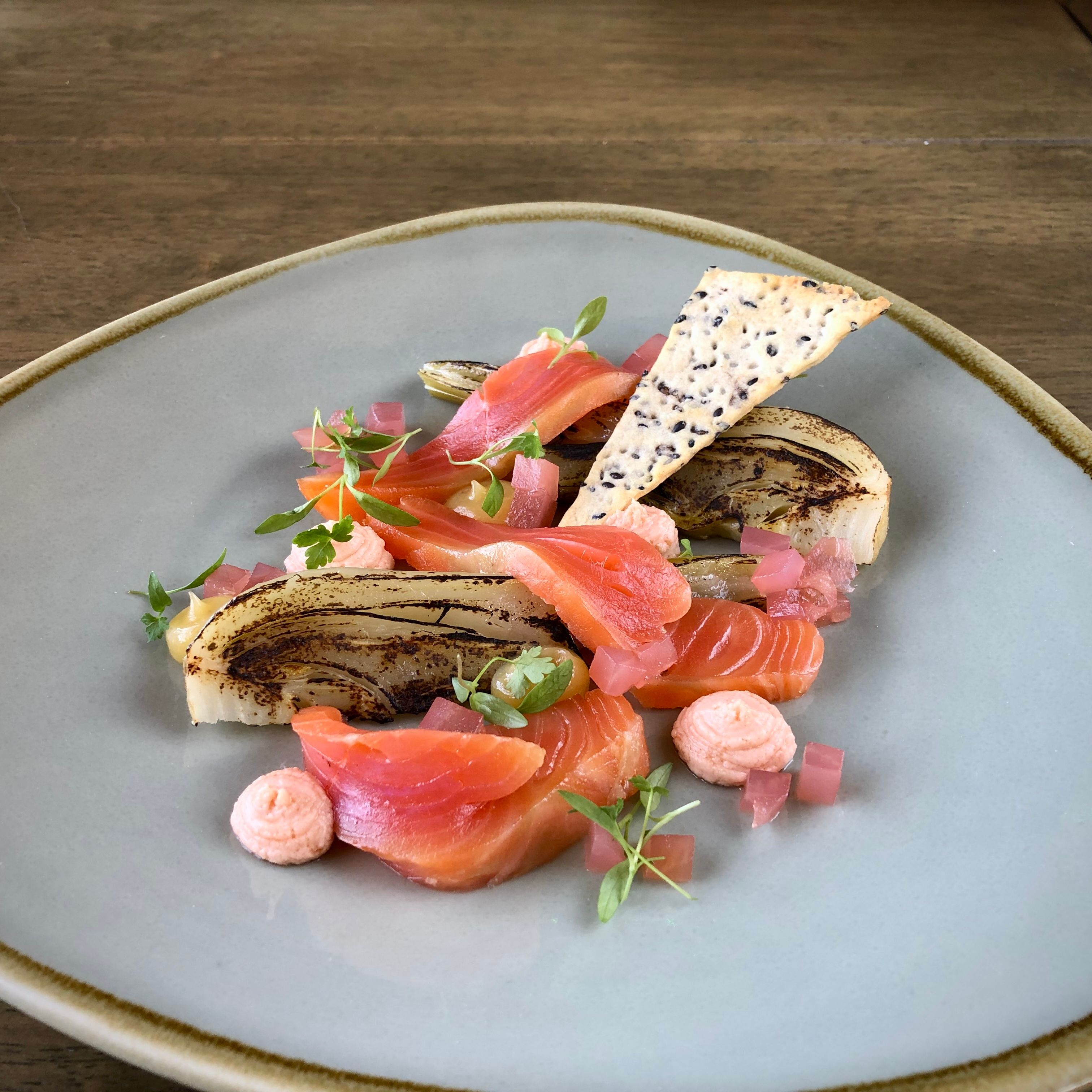 Gourmet plated dish with slices of cured salmon, grilled endive, pink garnish, microgreens, and a crisp cracker on a light grey plate.