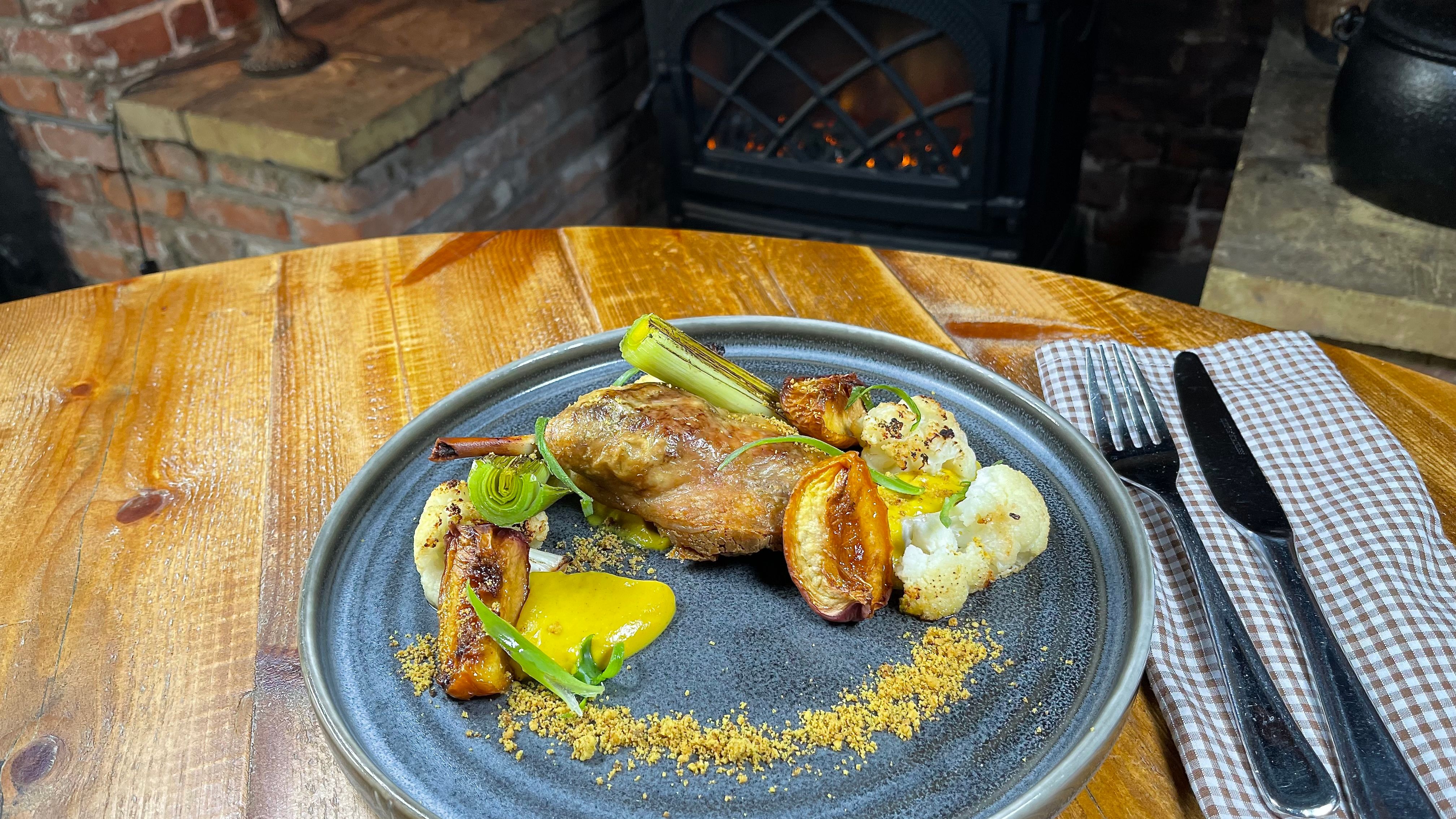 Gourmet plated dish with vegetables and meat on a rustic wooden table near a fireplace.
