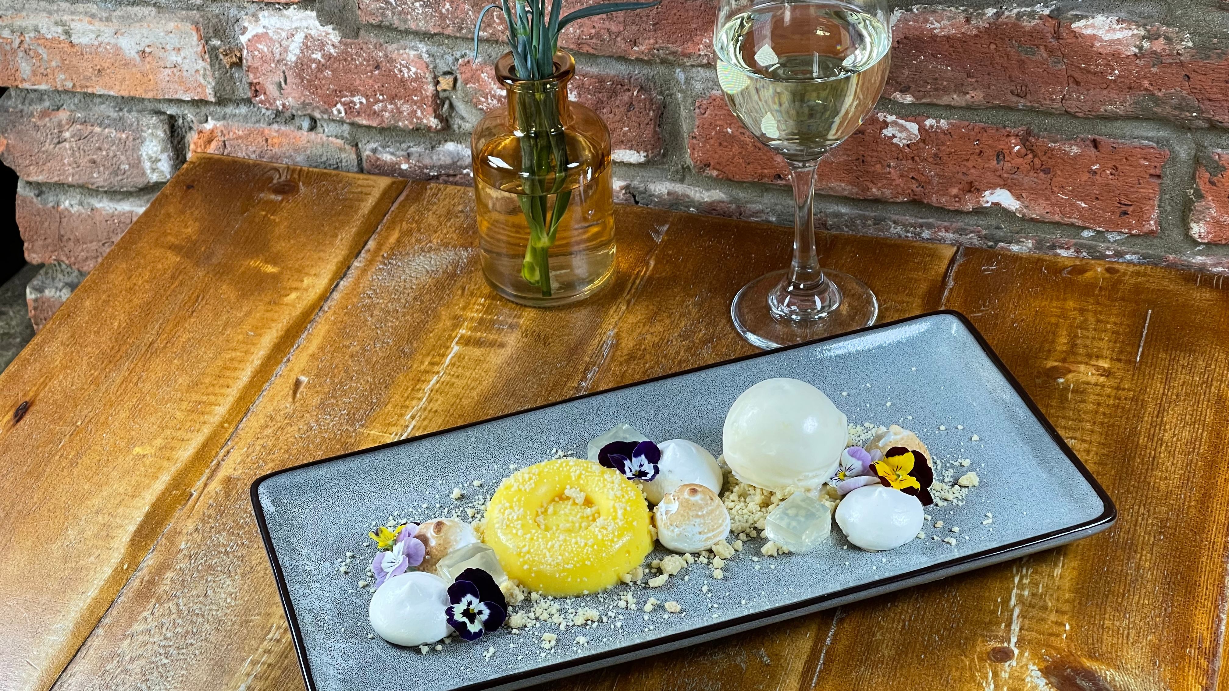 Elegant dessert plate garnished with flowers, glass of white wine, and a vase with a red carnation on a wooden table.