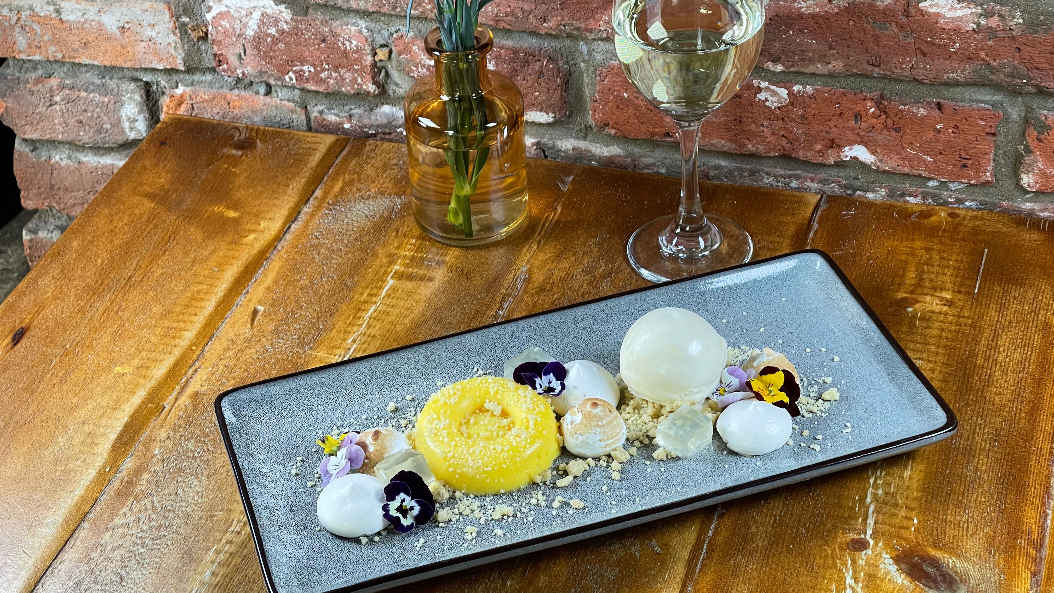 Elegant dessert plate garnished with flowers, glass of white wine, and a vase with a red carnation on a wooden table.