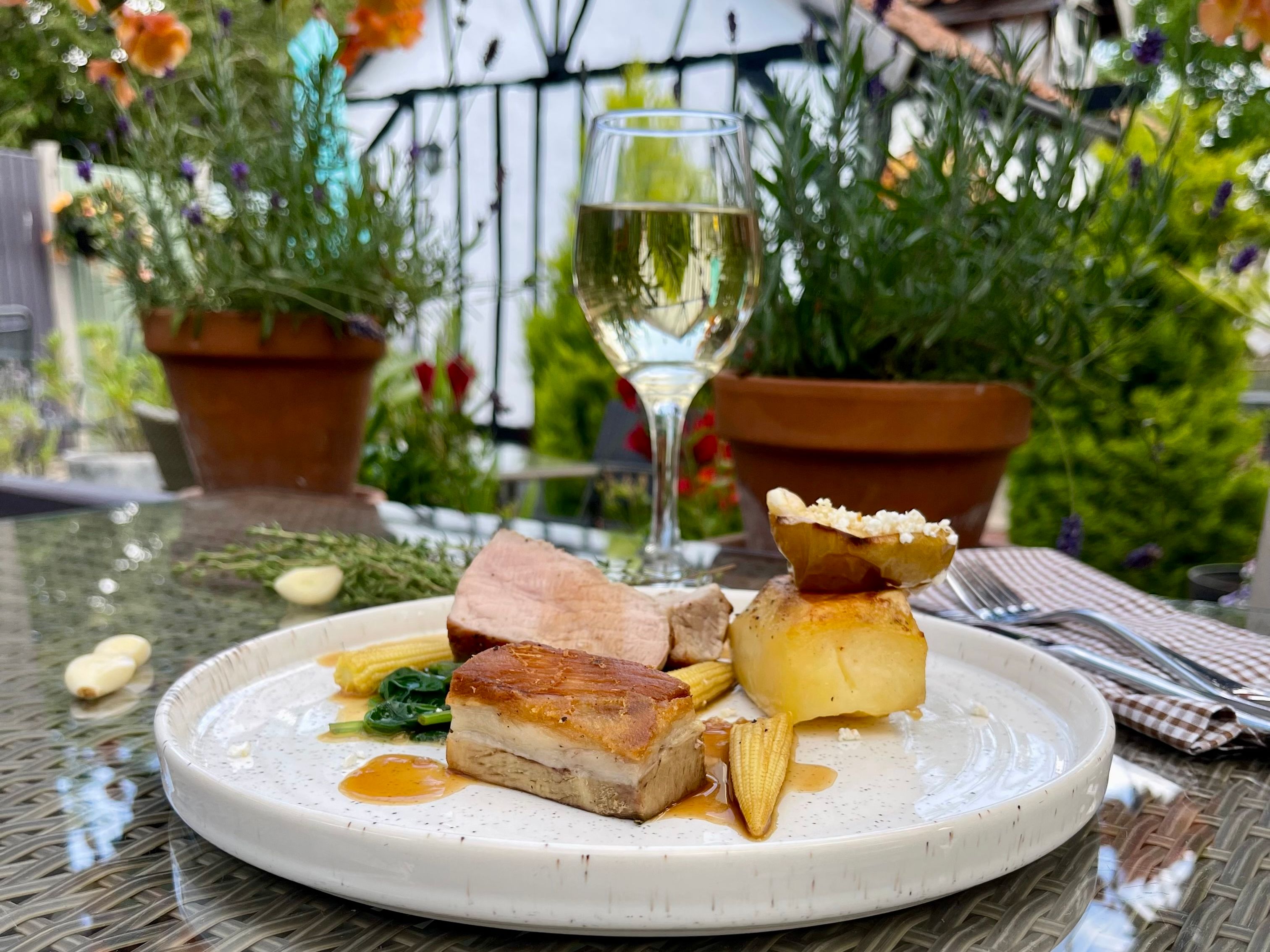 Gourmet plated meat dish with baby corn, greens and sauce on an outdoor table, with a glass of white wine behind