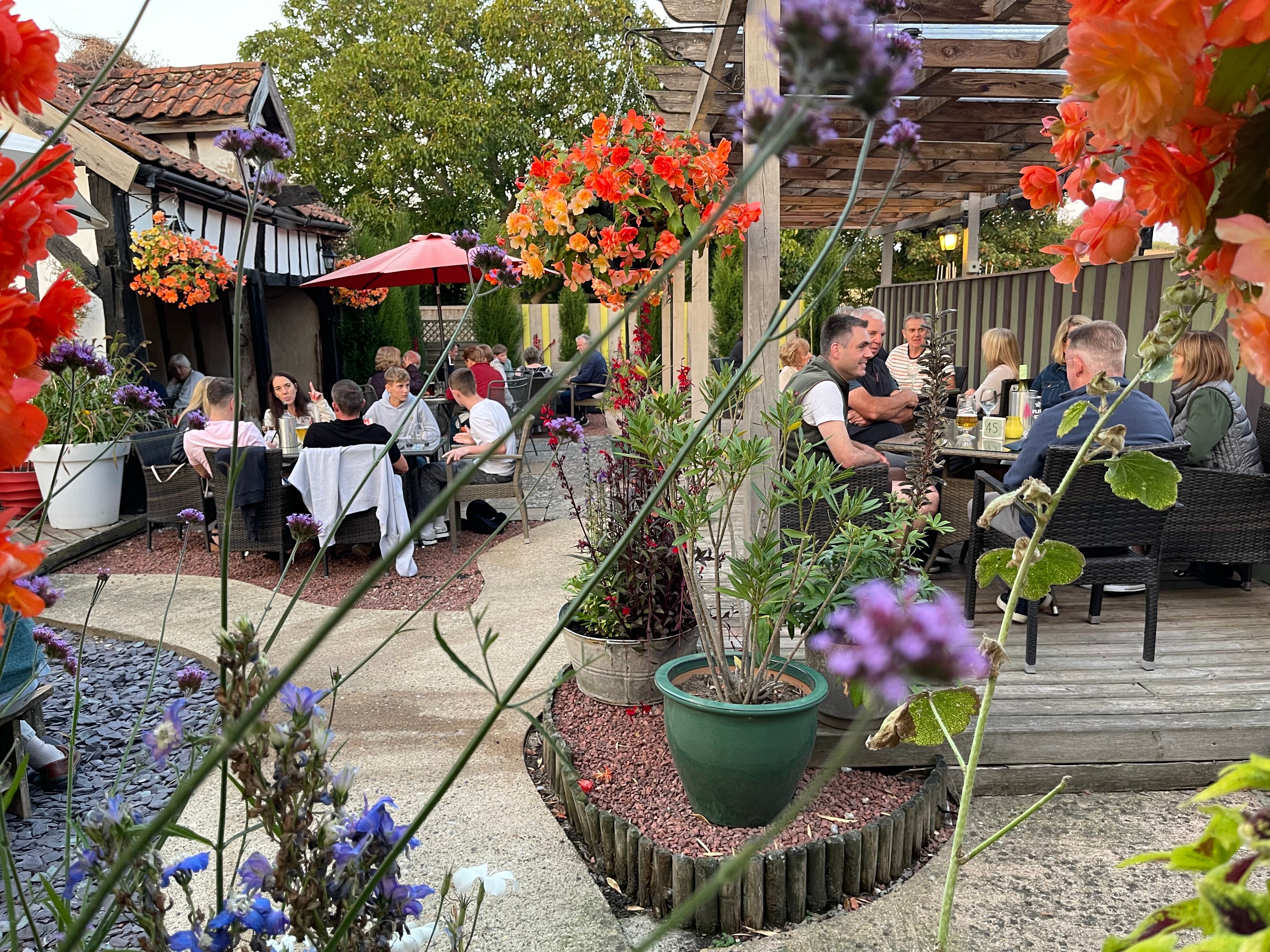 People dining outdoors in a garden courtyard with hanging baskets of bright flowers and a wooden pergola