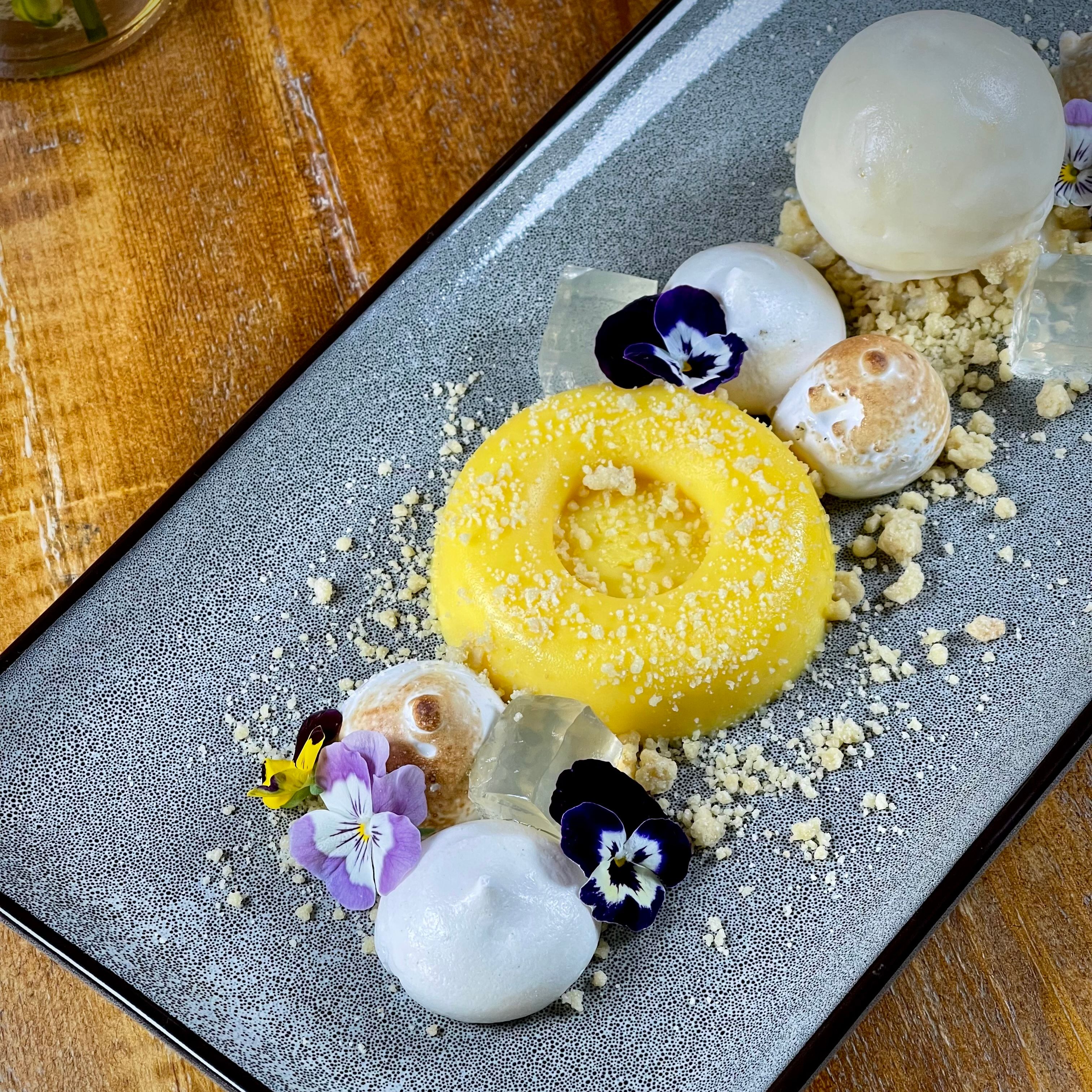 Elegant plated dessert featuring a yellow mousse ring, meringue drops, floral garnishes, and a white ice cream scoop on a gray rectangular plate.