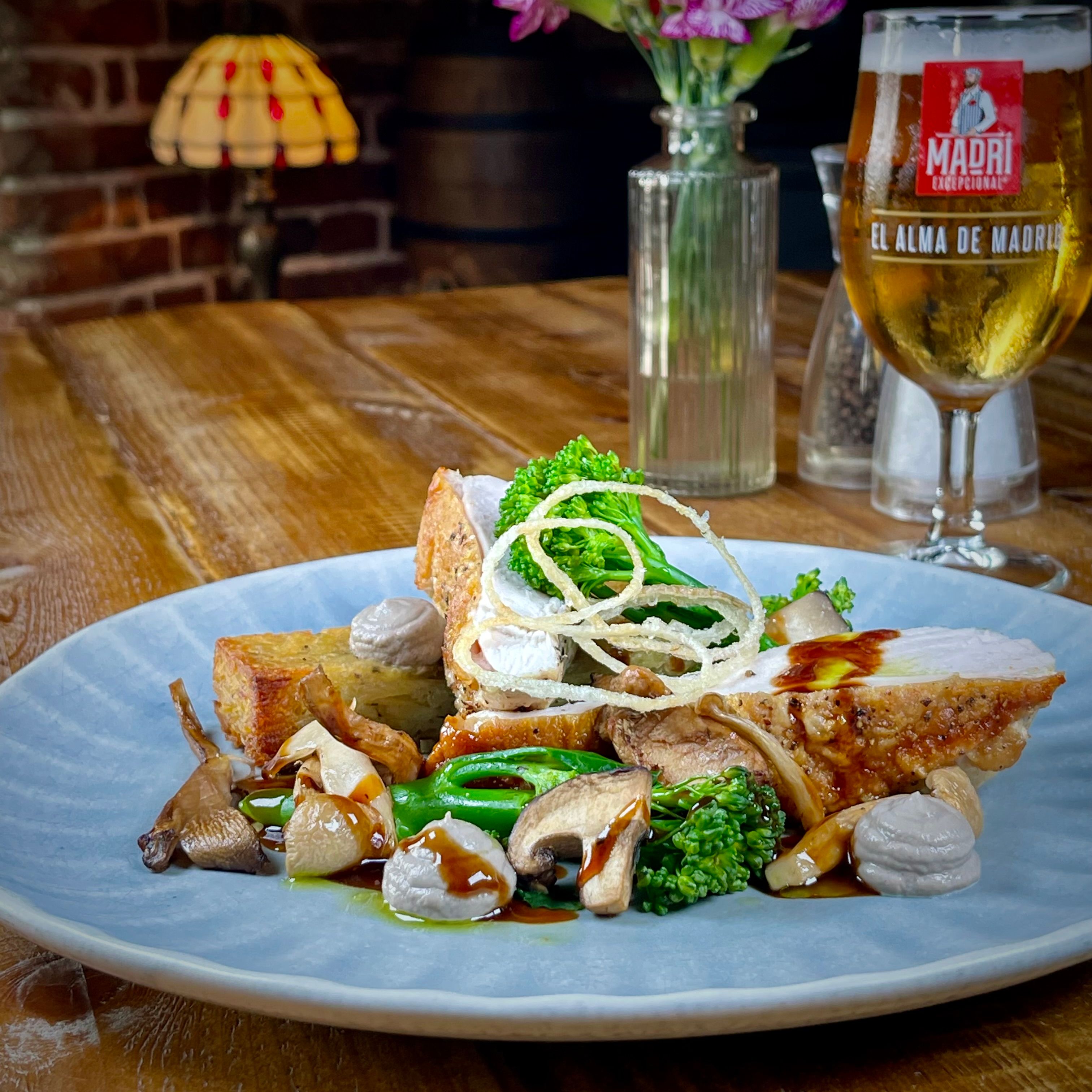 Gourmet chicken dish with vegetables, mushrooms, and crispy onions, served with a glass of beer on a wooden table