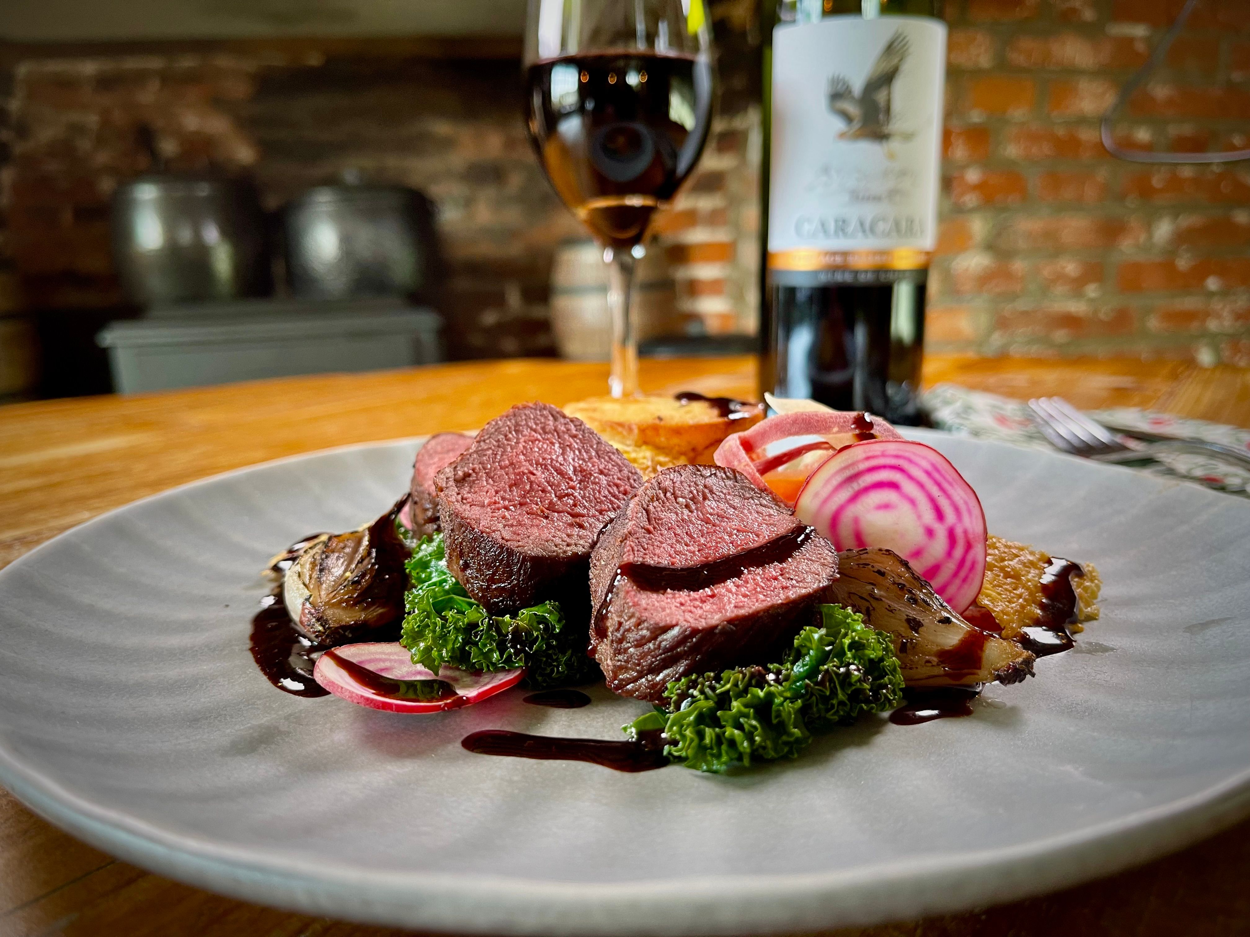 Plated gourmet meal featuring sliced venison, roasted vegetables, greens, and beet slices, with a glass and bottle of red wine in the background.