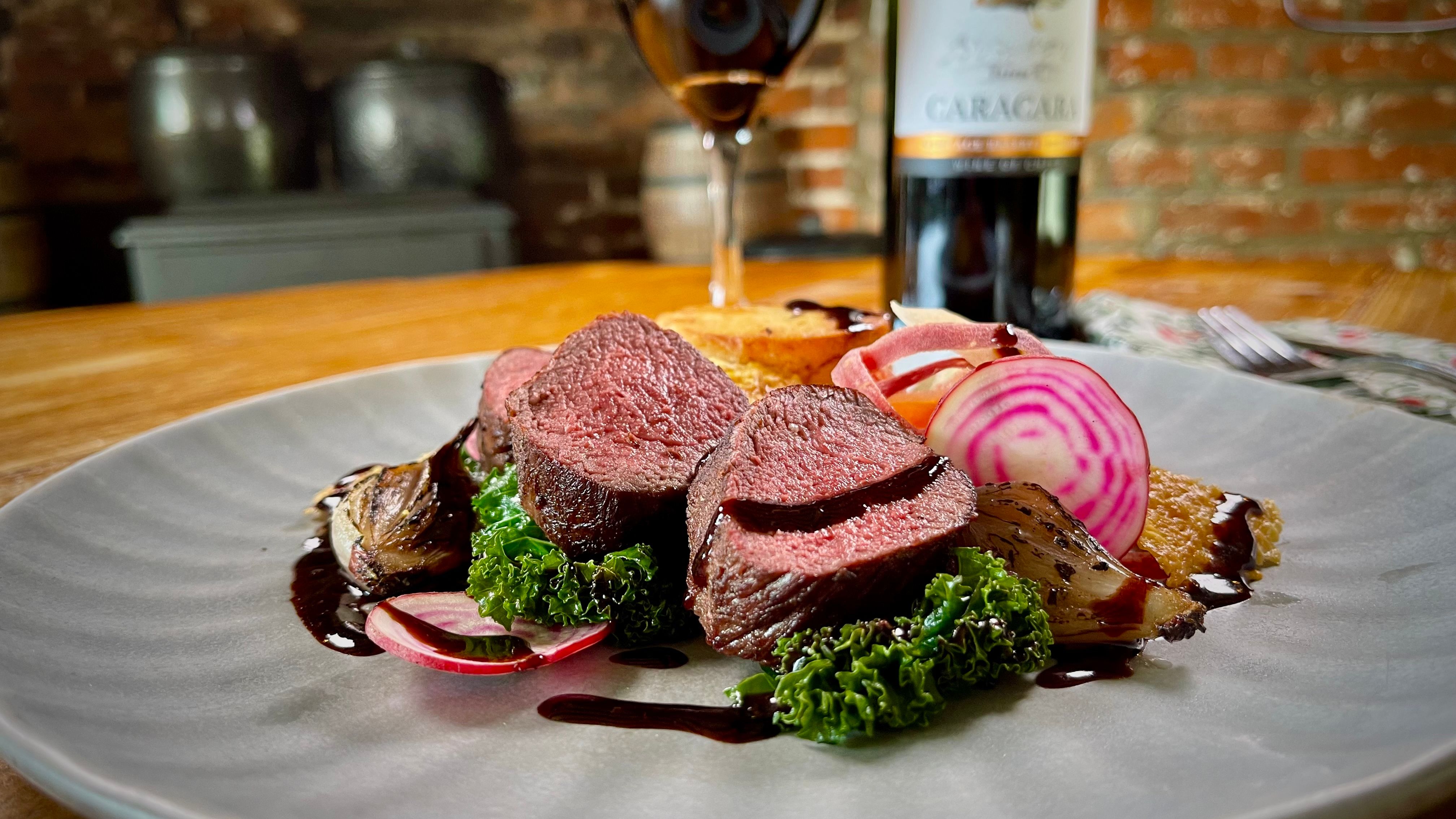Plated gourmet meal featuring sliced venison, roasted vegetables, greens, and beet slices, with a glass and bottle of red wine in the background.