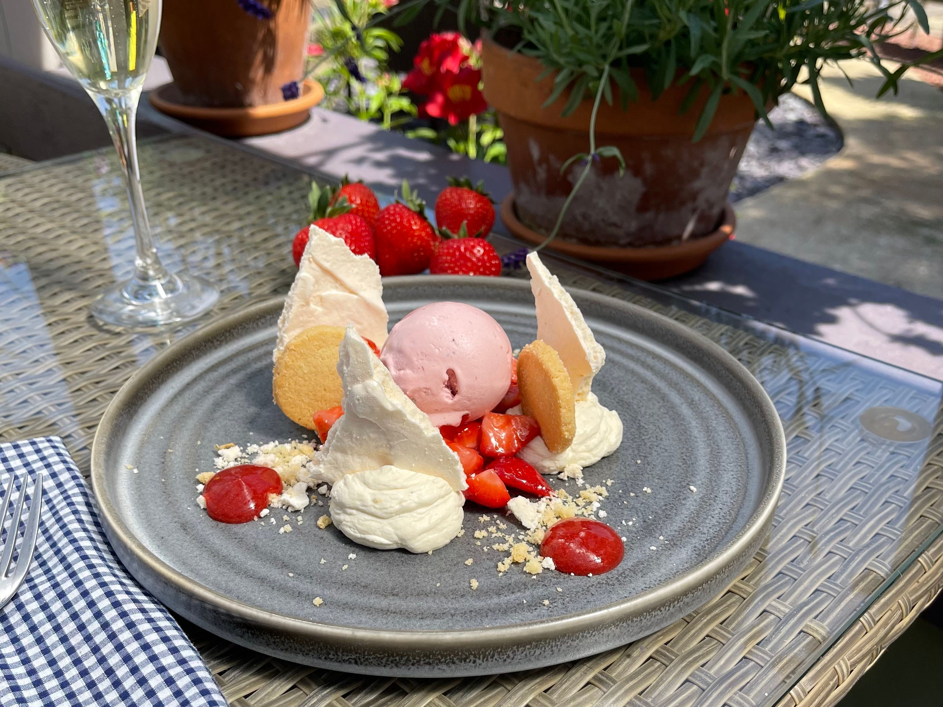 A dessert plate with strawberry ice cream, meringue, whipped cream, fresh strawberries, and cookies, next to a glass of sparkling wine on an outdoor table.