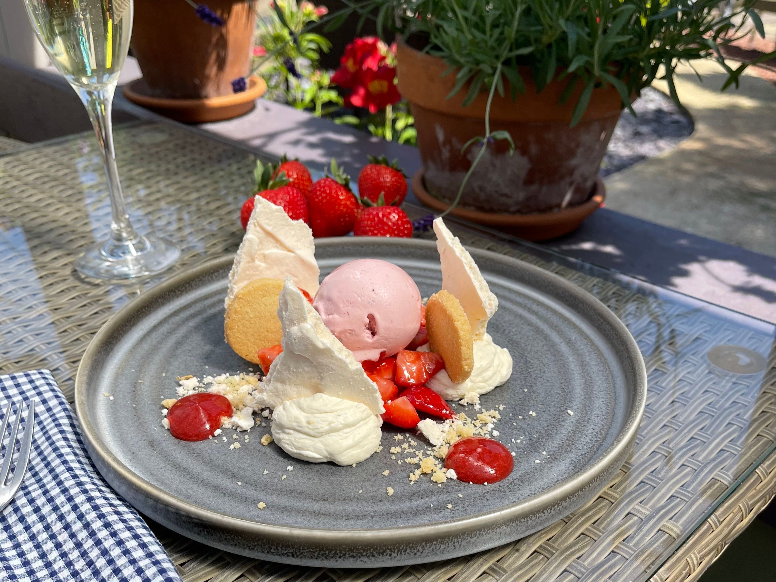 A dessert plate with strawberry ice cream, meringue, whipped cream, fresh strawberries, and cookies, next to a glass of sparkling wine on an outdoor table.