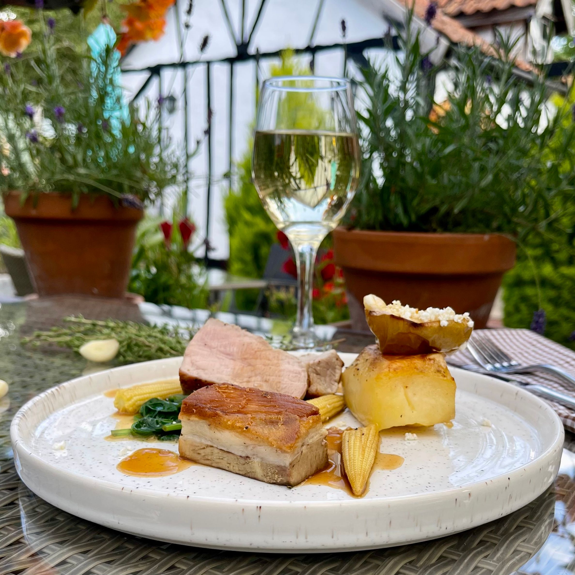 Gourmet meal with roast pork, potato, baby corn, and a glass of white wine on an outdoor table with plants in the background