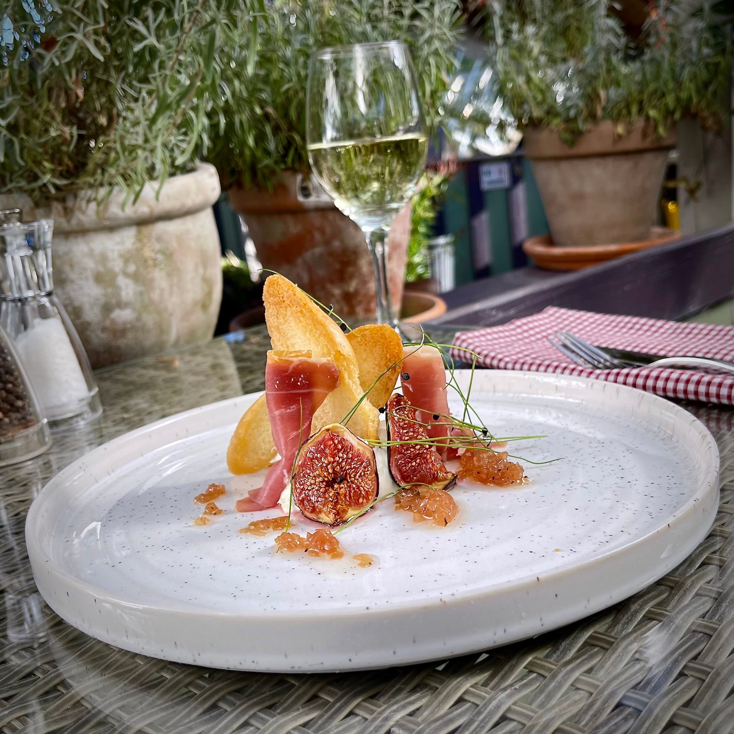 A gourmet dish with figs, prosciutto, toasted bread, and garnishes on a white plate beside a glass of white wine. Outdoor table setting with potted plants in the background.