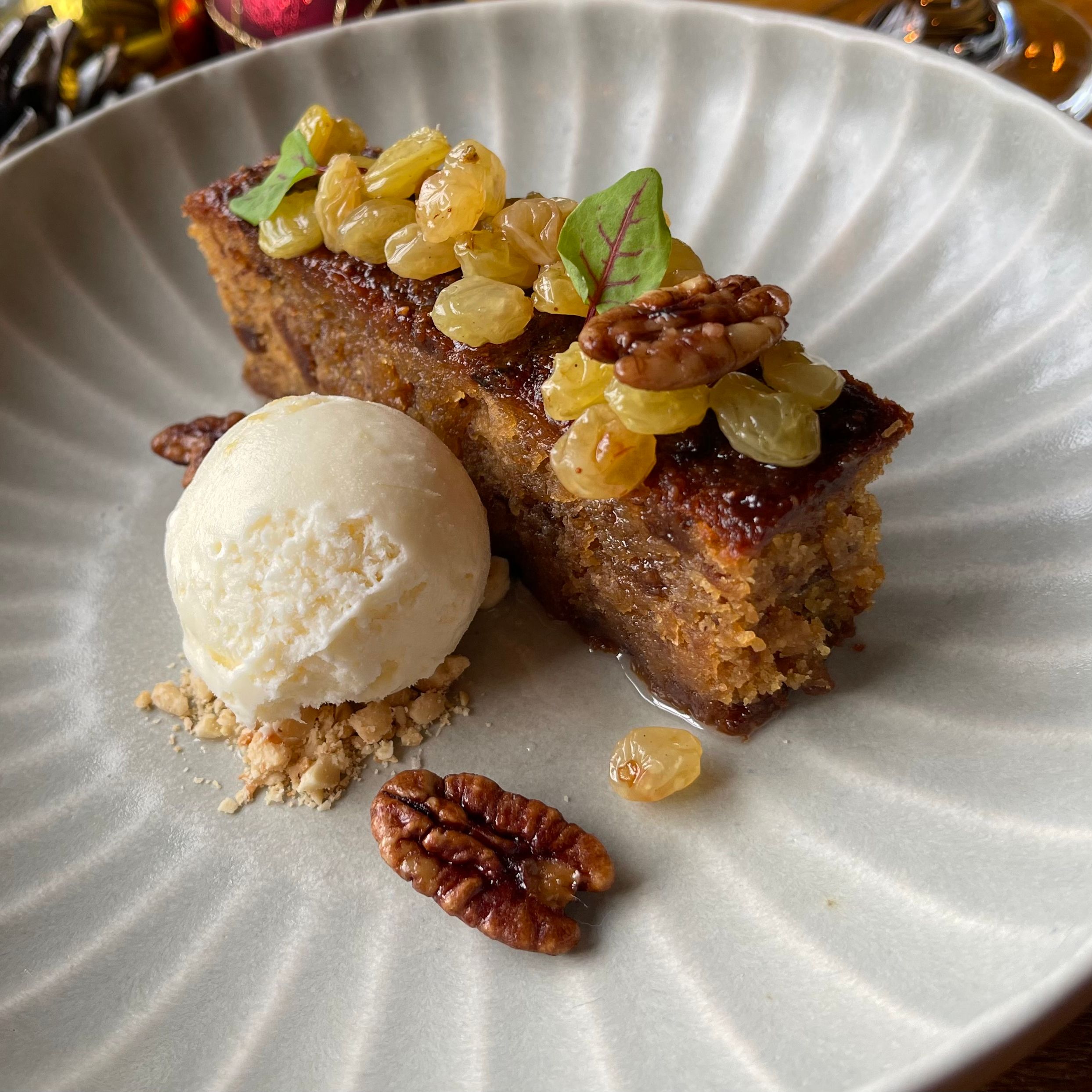 A plated dessert featuring a slice of cake topped with golden raisins and pecans, served with a scoop of vanilla ice cream.
