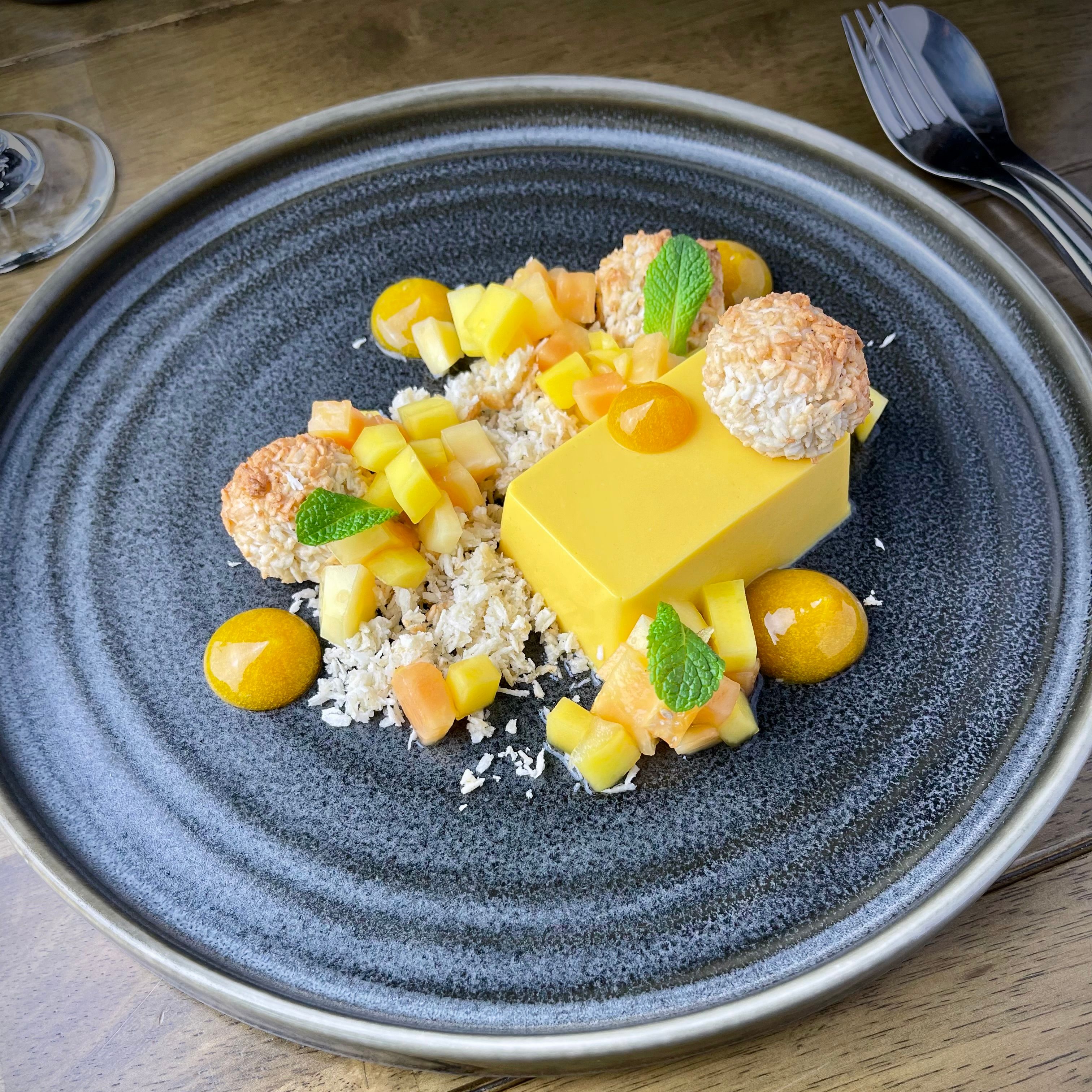 Plated mango dessert with diced fruit, coconut balls, and mint leaves on a dark ceramic plate
