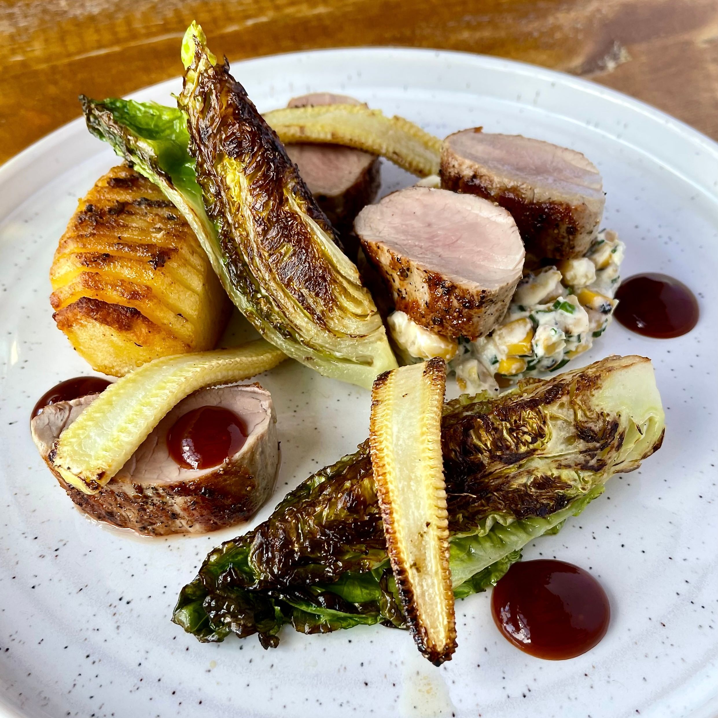 Plated gourmet dish with sliced pork tenderloin, baby corn, roasted potato, charred lettuce, and sauce droplets
