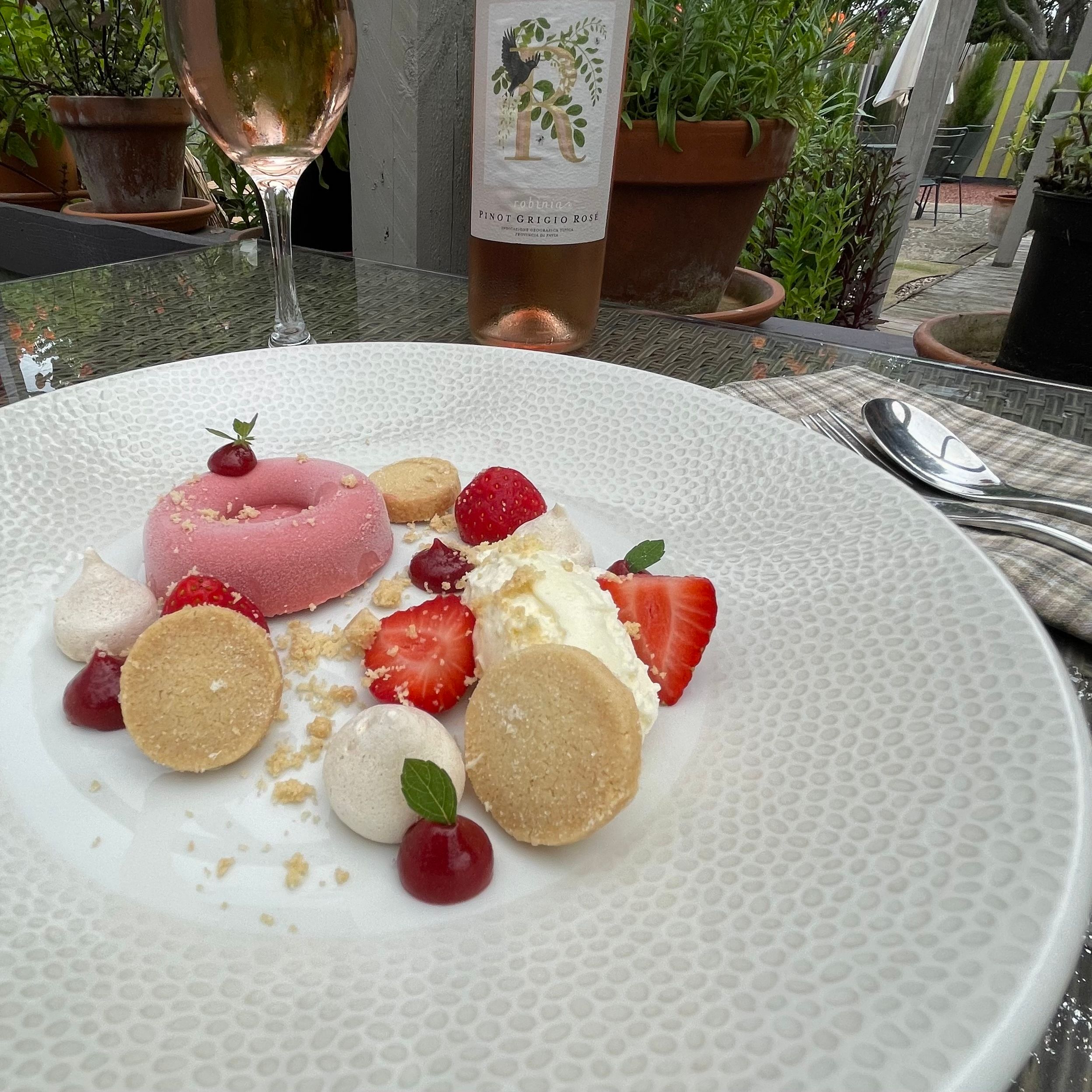 A plate of elegant strawberry dessert with a glass and bottle of rosé wine on an outdoor table surrounded by flowers.