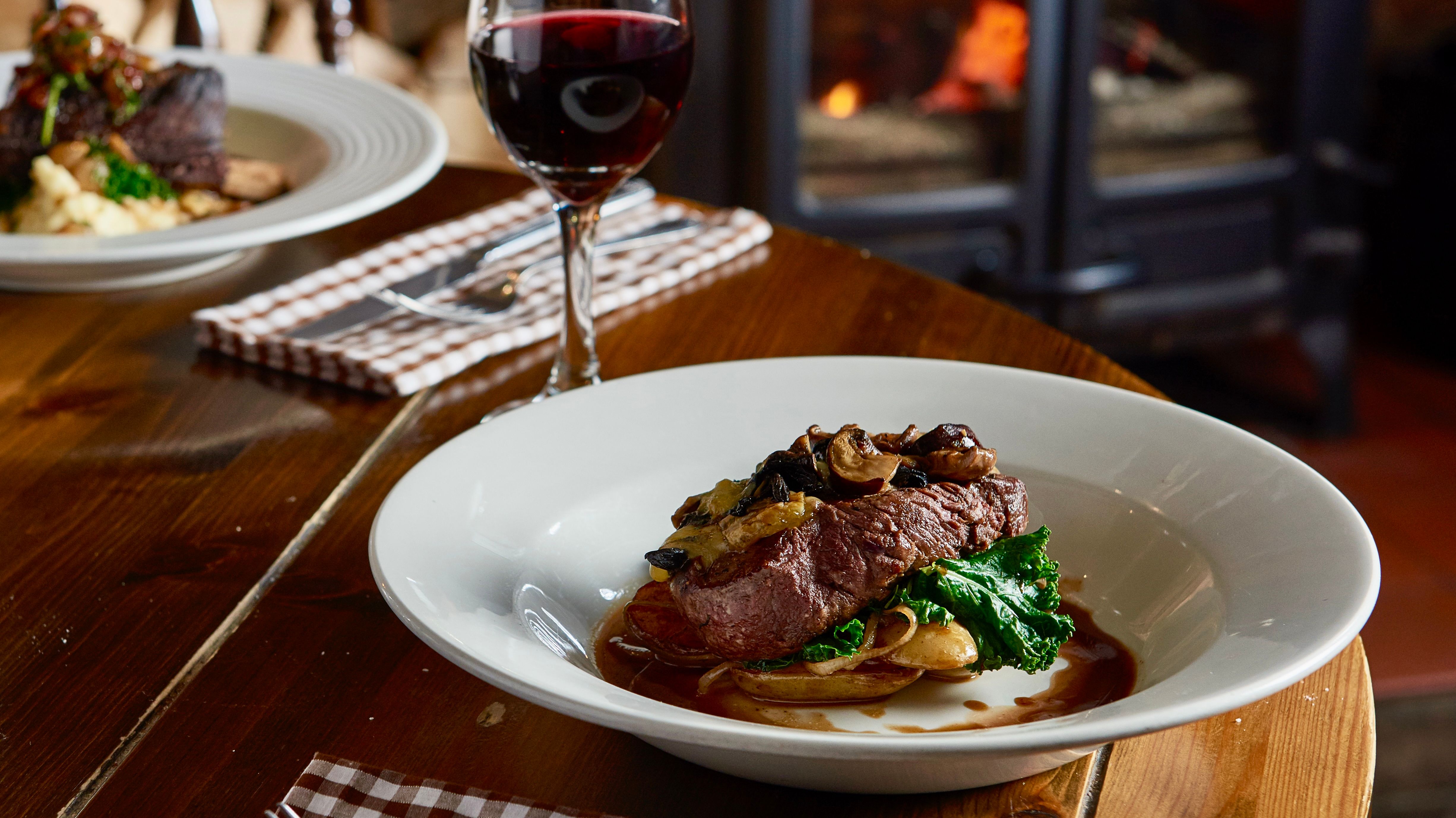 Steak dinner with vegetables and mushrooms served with a glass of red wine in a cozy restaurant with a wood-burning stove in the background.