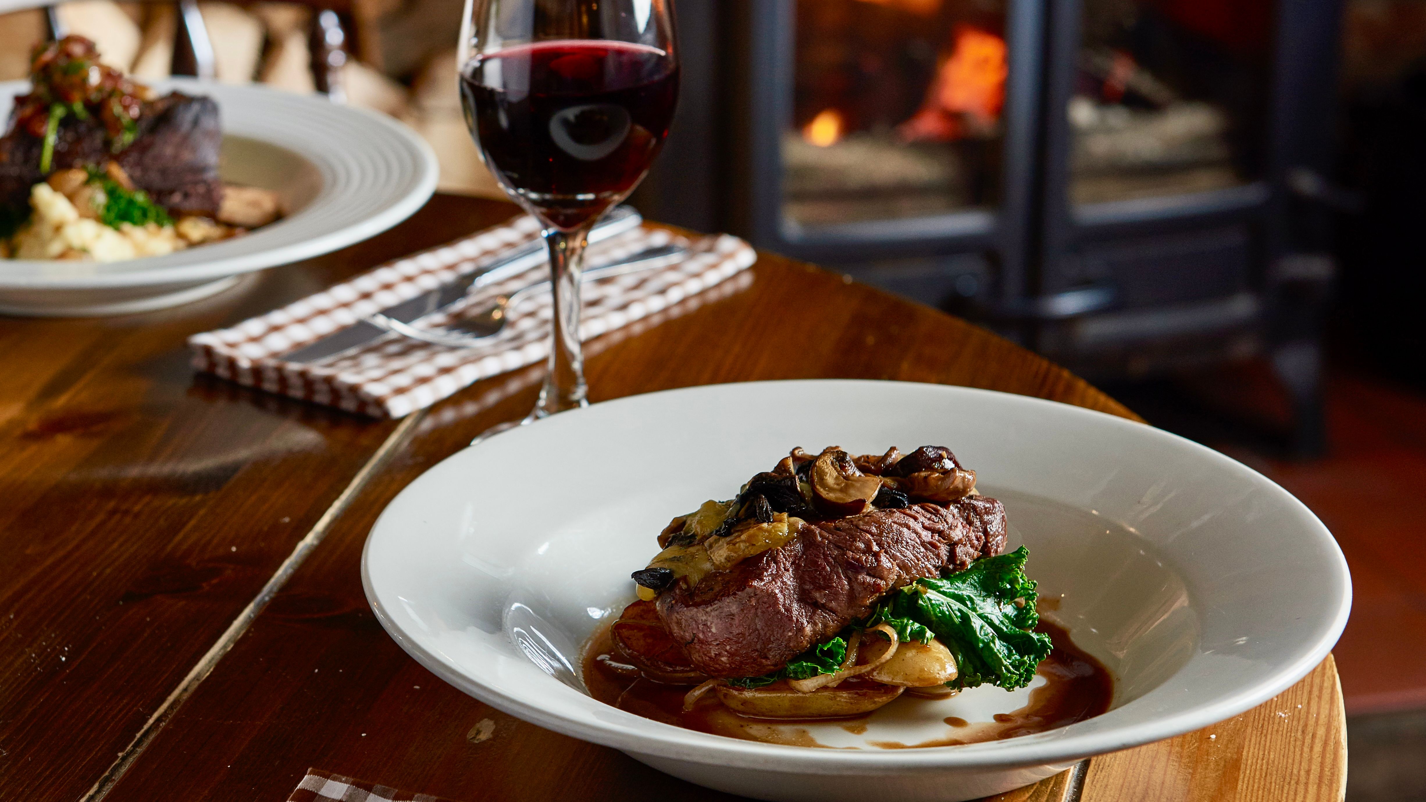 Steak dinner with vegetables and mushrooms served with a glass of red wine in a cozy restaurant with a wood-burning stove in the background.