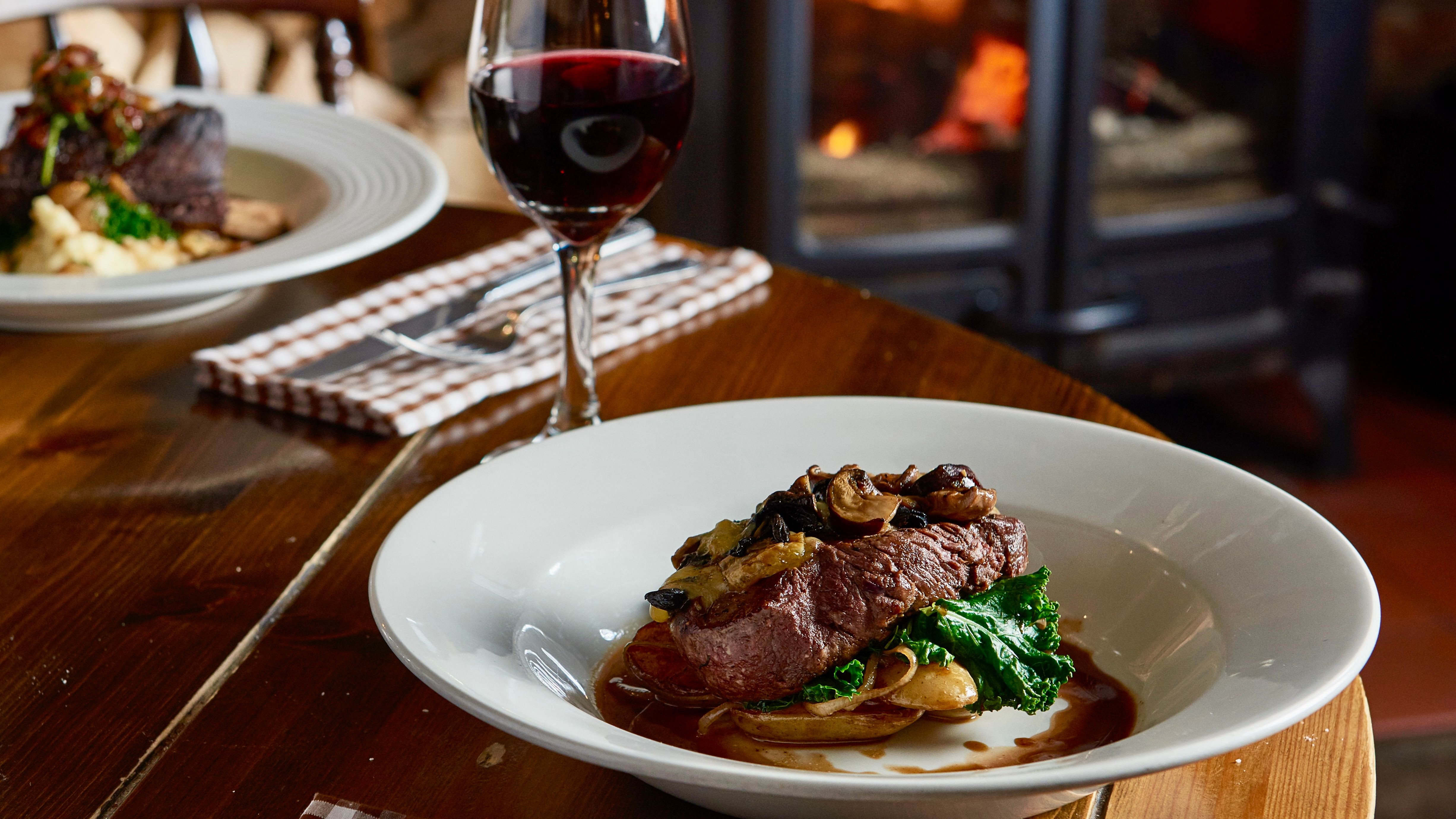 Steak dinner with vegetables and mushrooms served with a glass of red wine in a cozy restaurant with a wood-burning stove in the background.