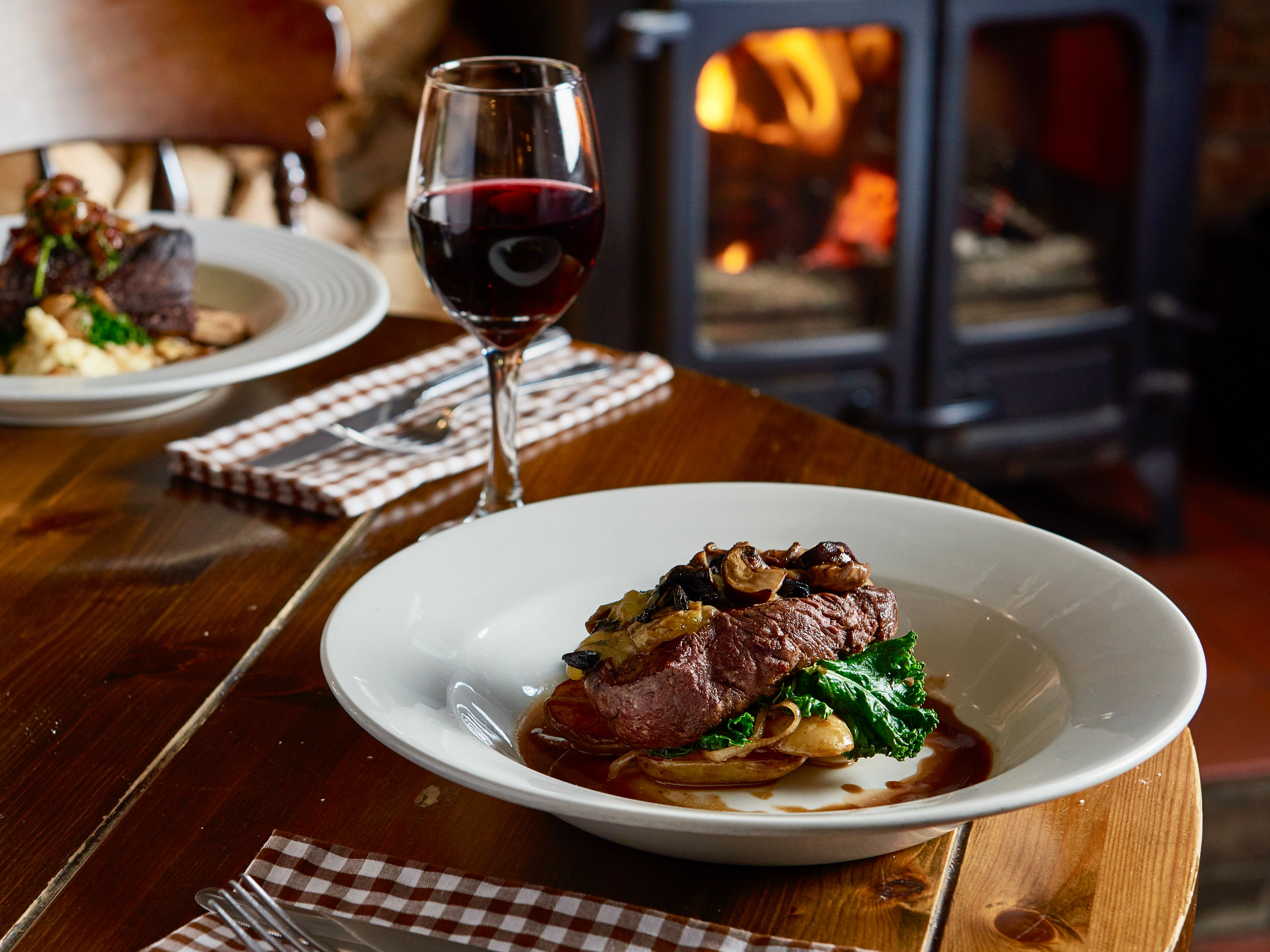 Steak dinner with vegetables and mushrooms served with a glass of red wine in a cozy restaurant with a wood-burning stove in the background.