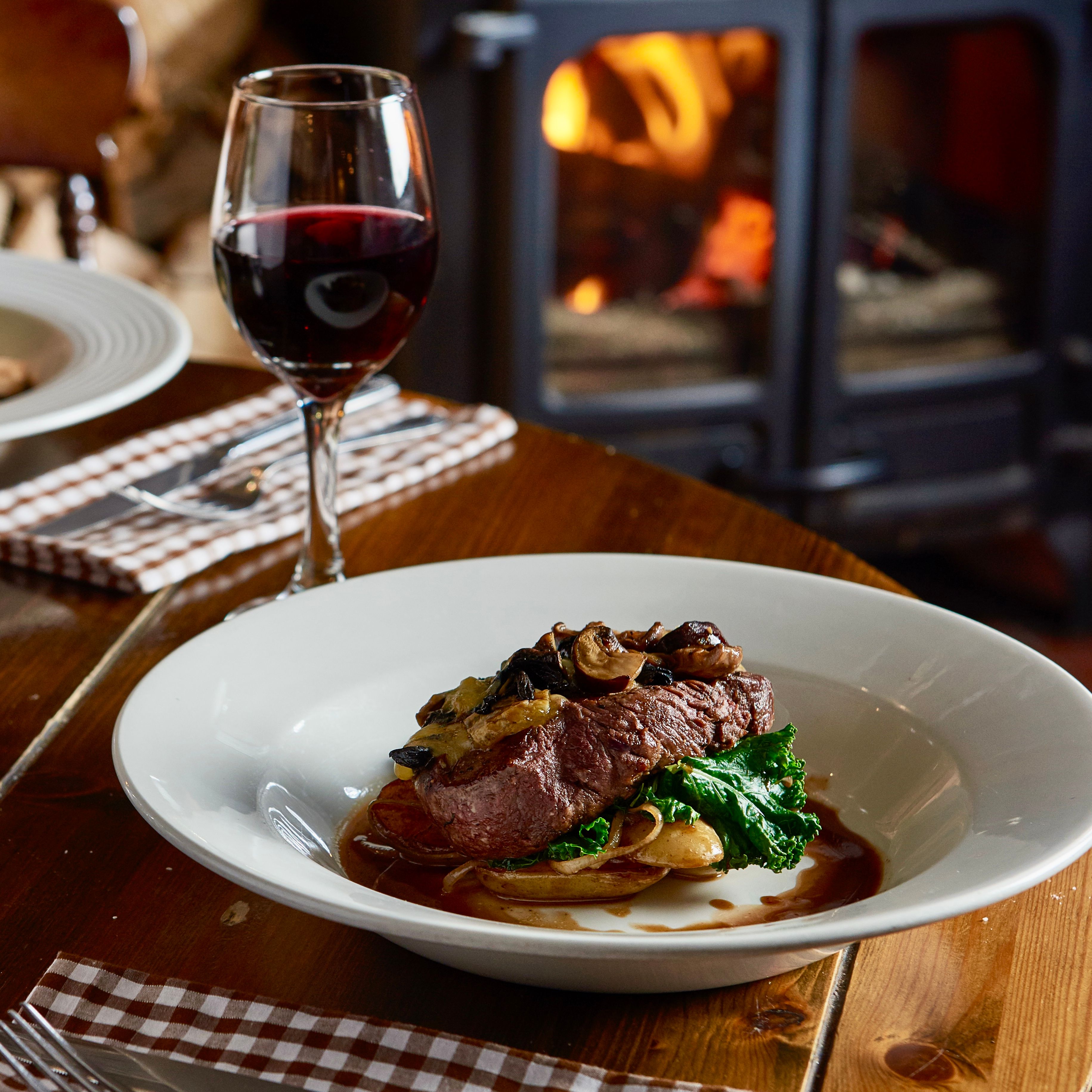 Steak dinner with vegetables and mushrooms served with a glass of red wine in a cozy restaurant with a wood-burning stove in the background.
