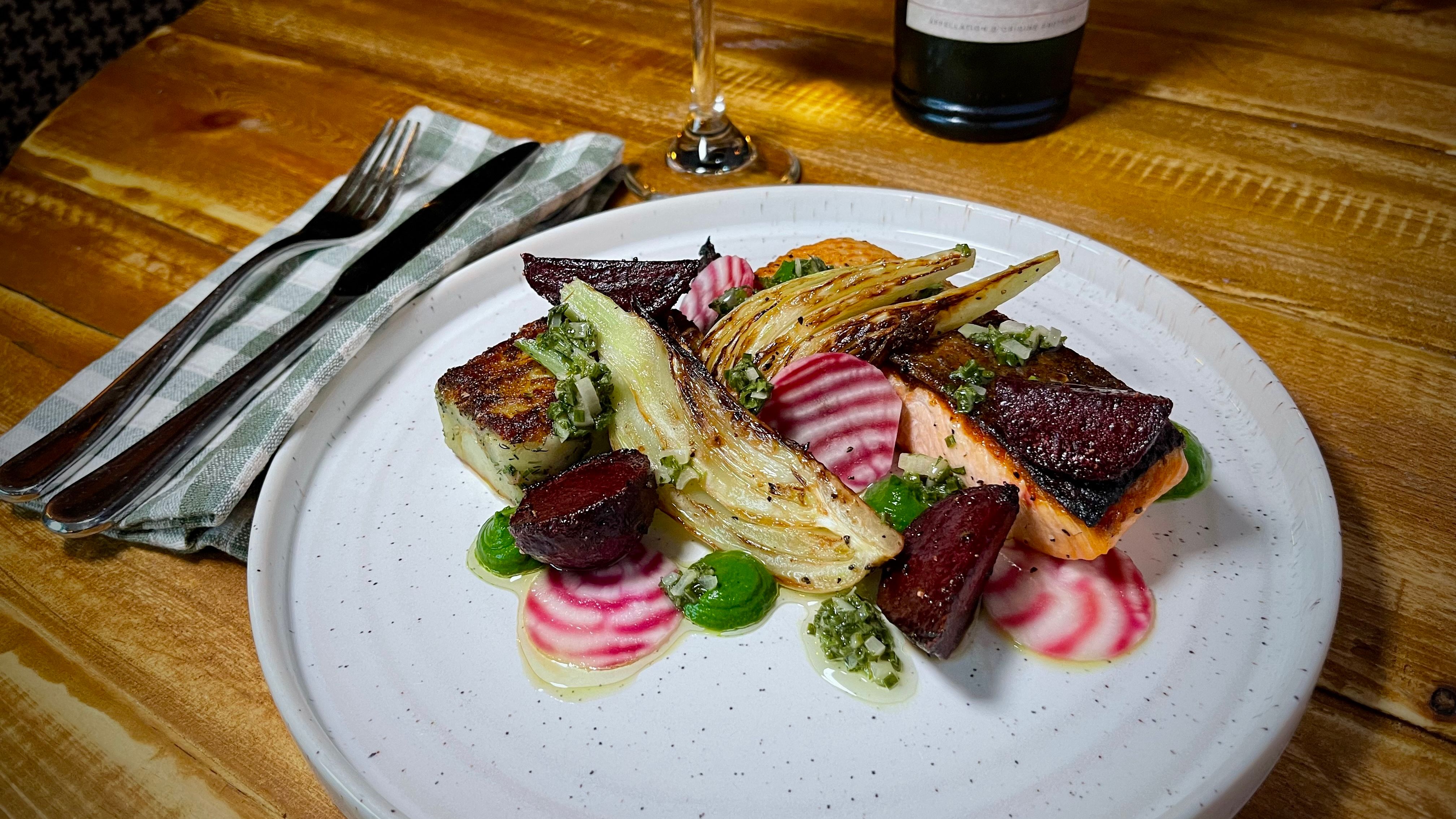 Gourmet salmon dish with vegetables on a white plate next to a wine glass and bottle