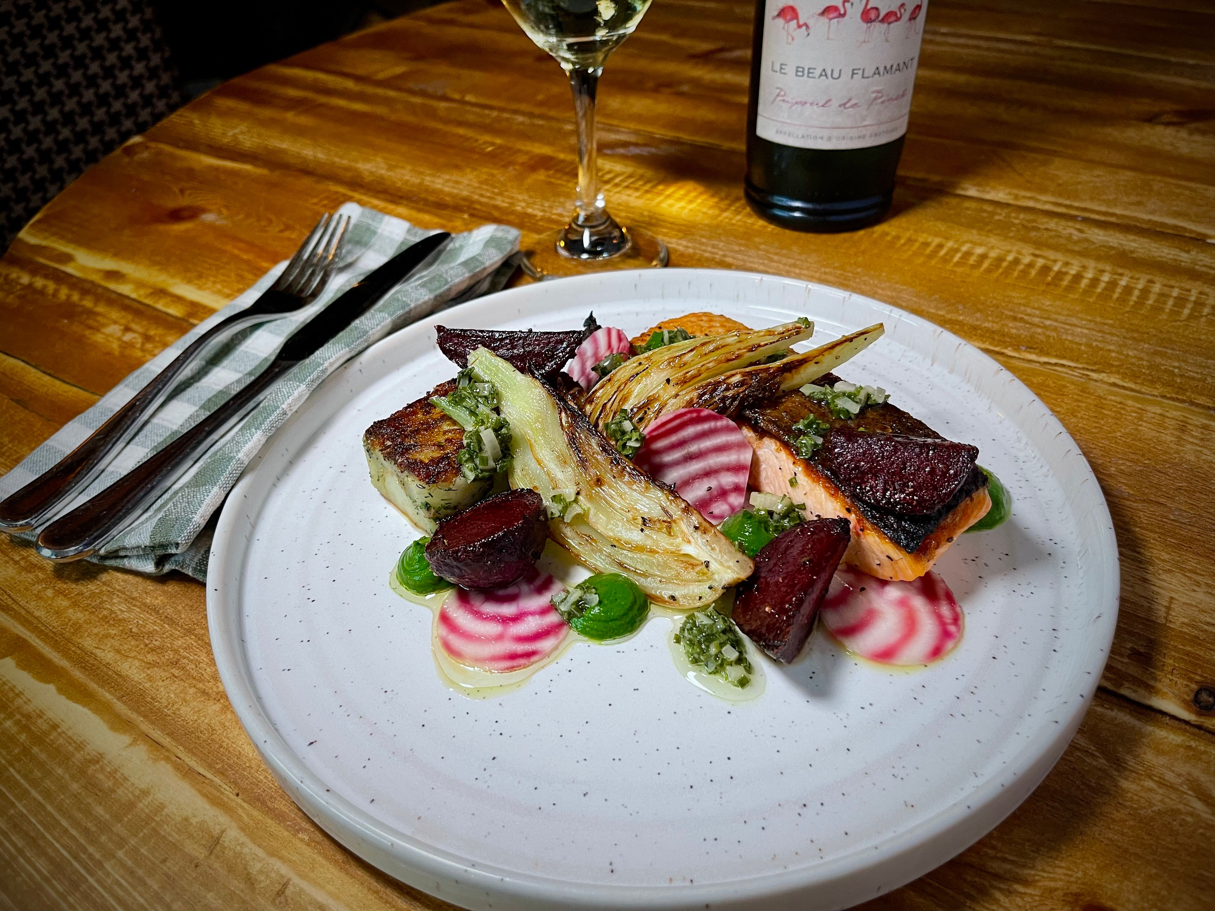 Gourmet salmon dish with vegetables on a white plate next to a wine glass and bottle