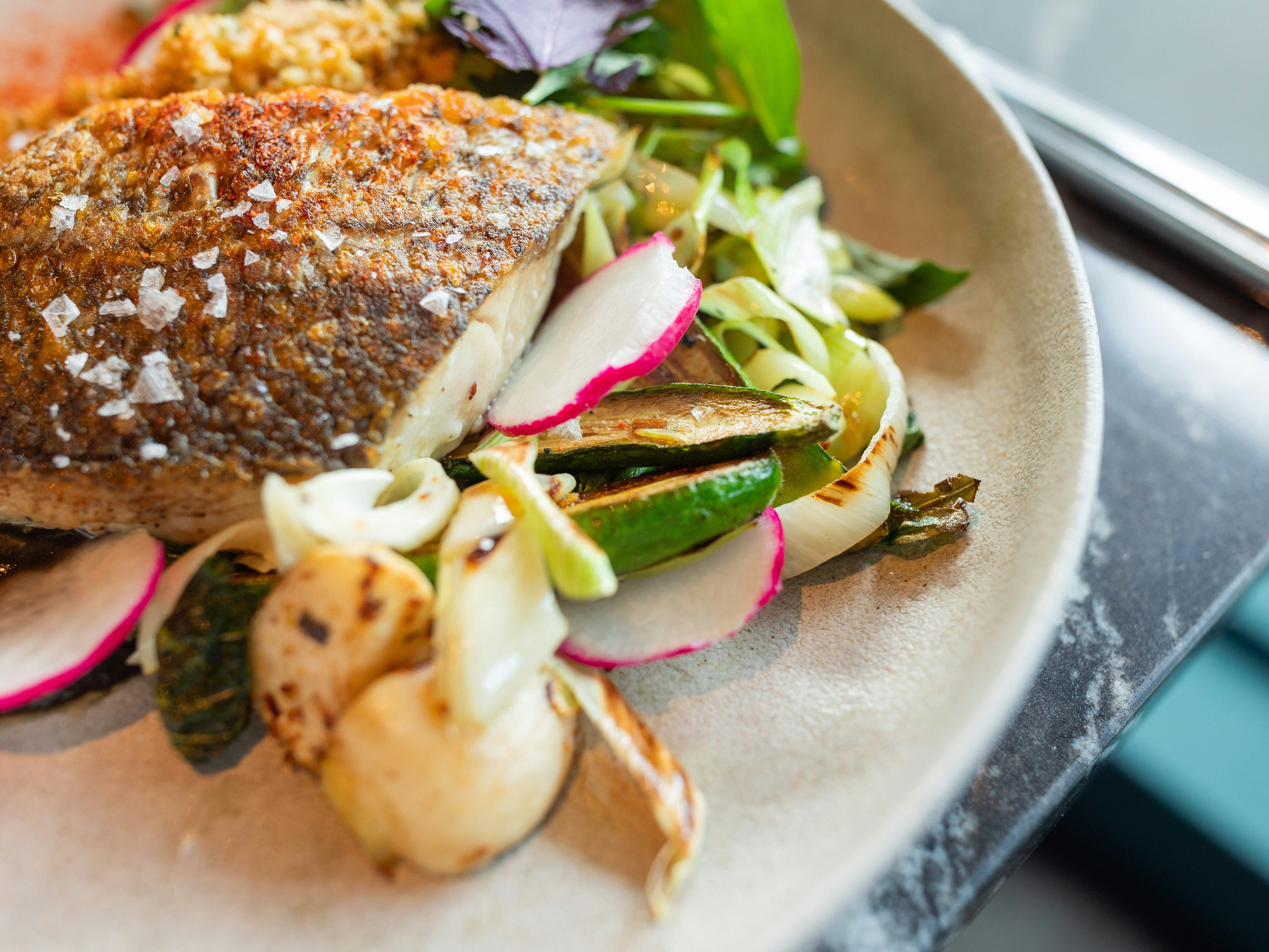 Grilled fish fillet served with a side of sautéed vegetables including radishes, zucchini, and onions, garnished with flakes of sea salt.