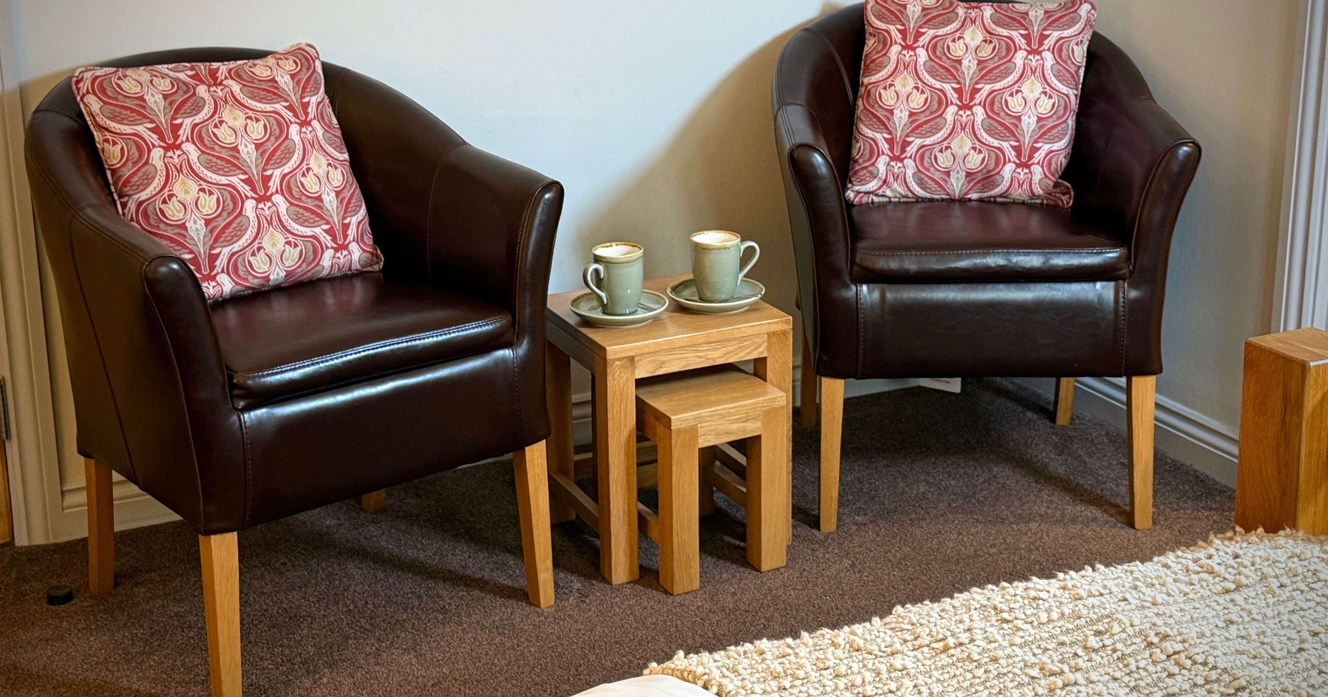 Two brown leather armchairs with red patterned cushions and a small wooden nesting table set between them holding two cups of coffee.