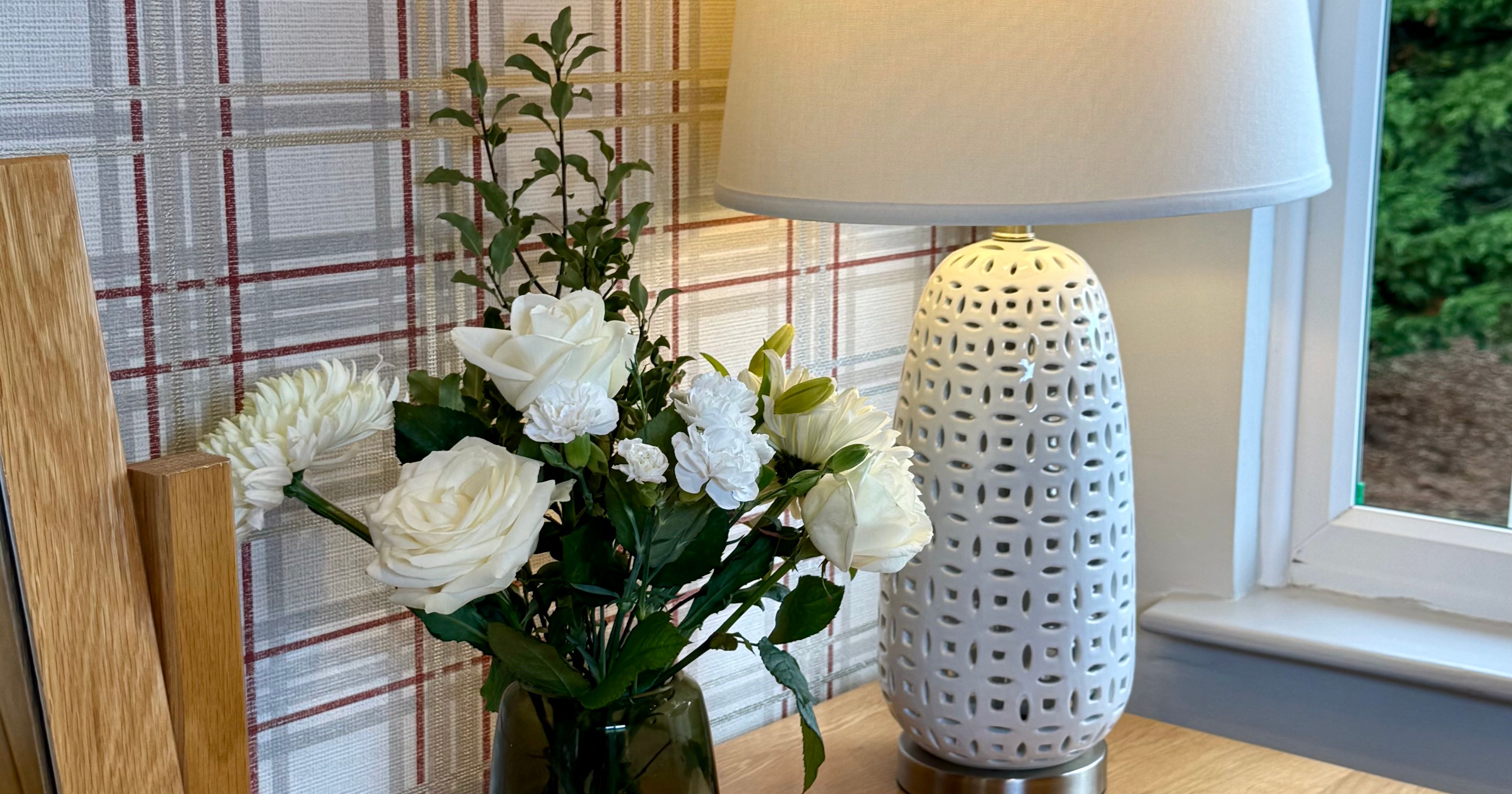 A white ceramic table lamp and a vase of white flowers on a wooden desk by a window.