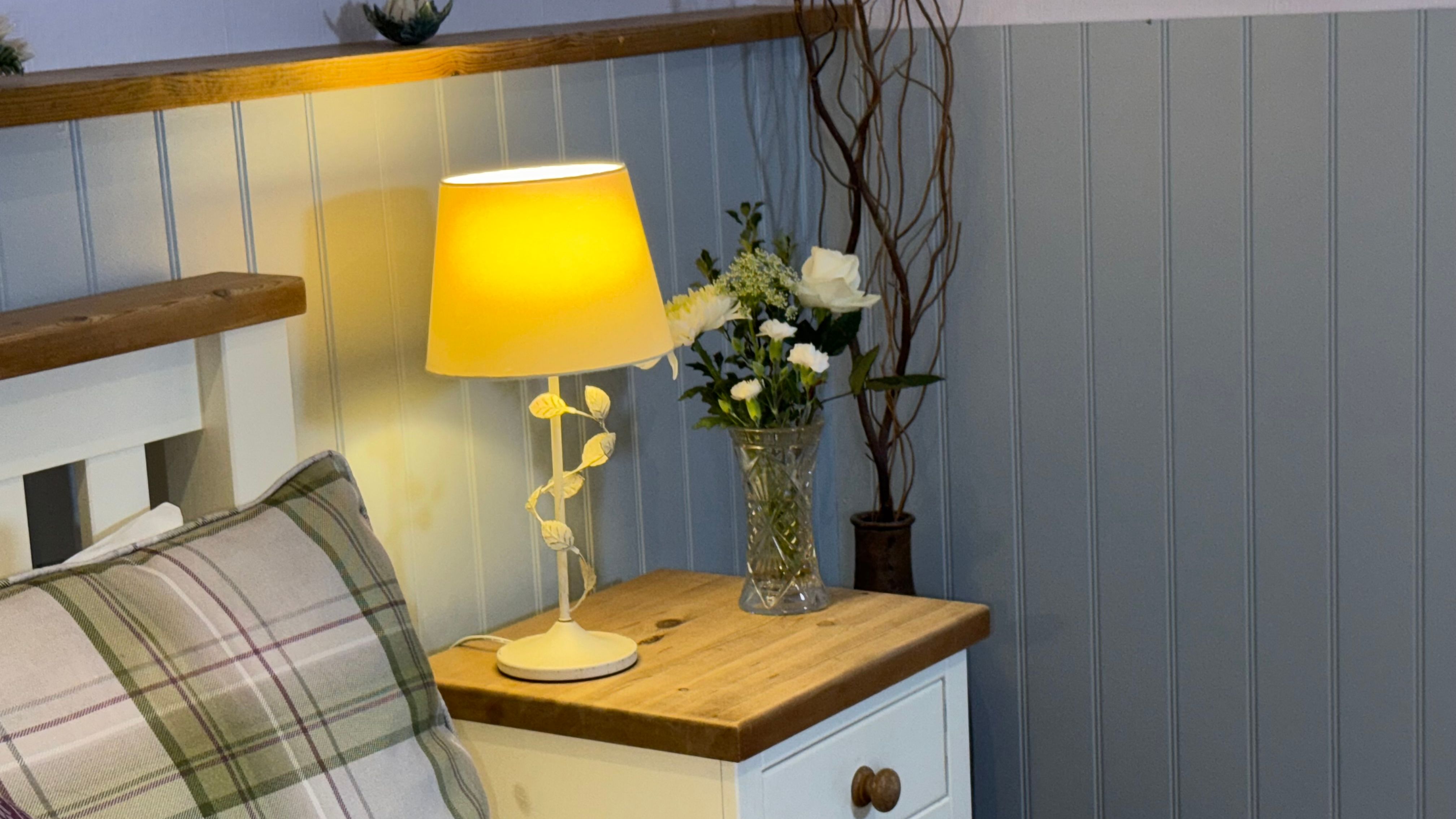 Bedside table with a lamp, flowers in a vase, and a decorative pillow.