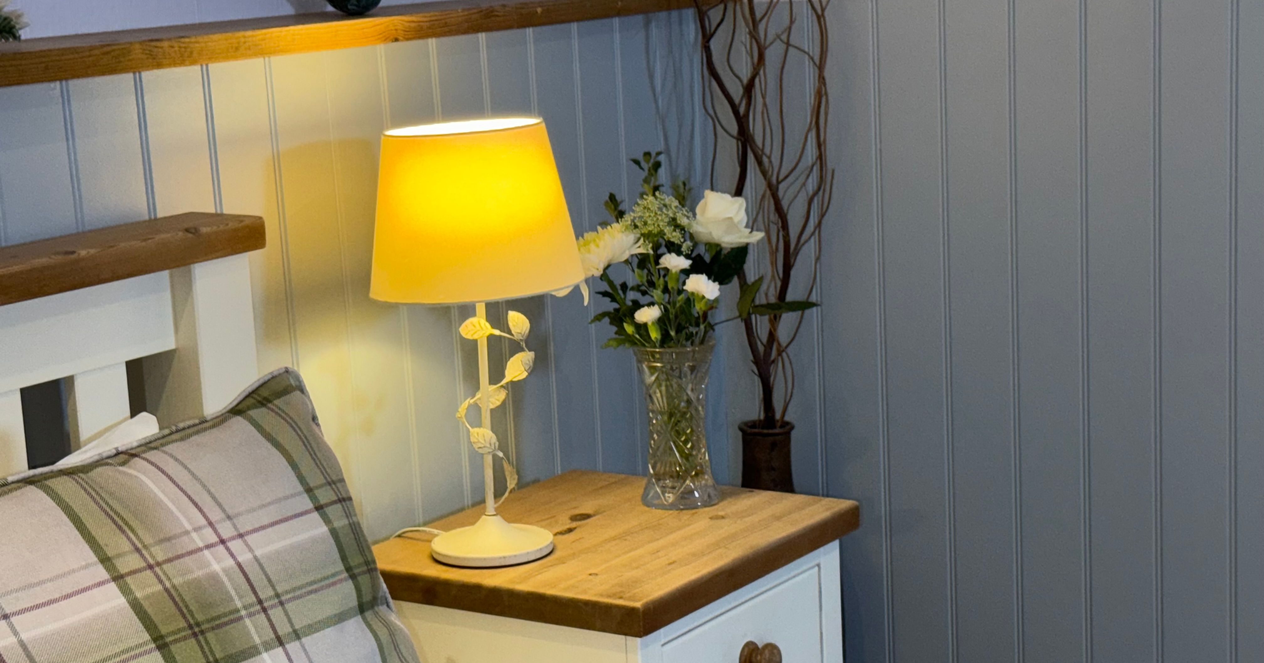 Bedside table with a lamp, flowers in a vase, and a decorative pillow.