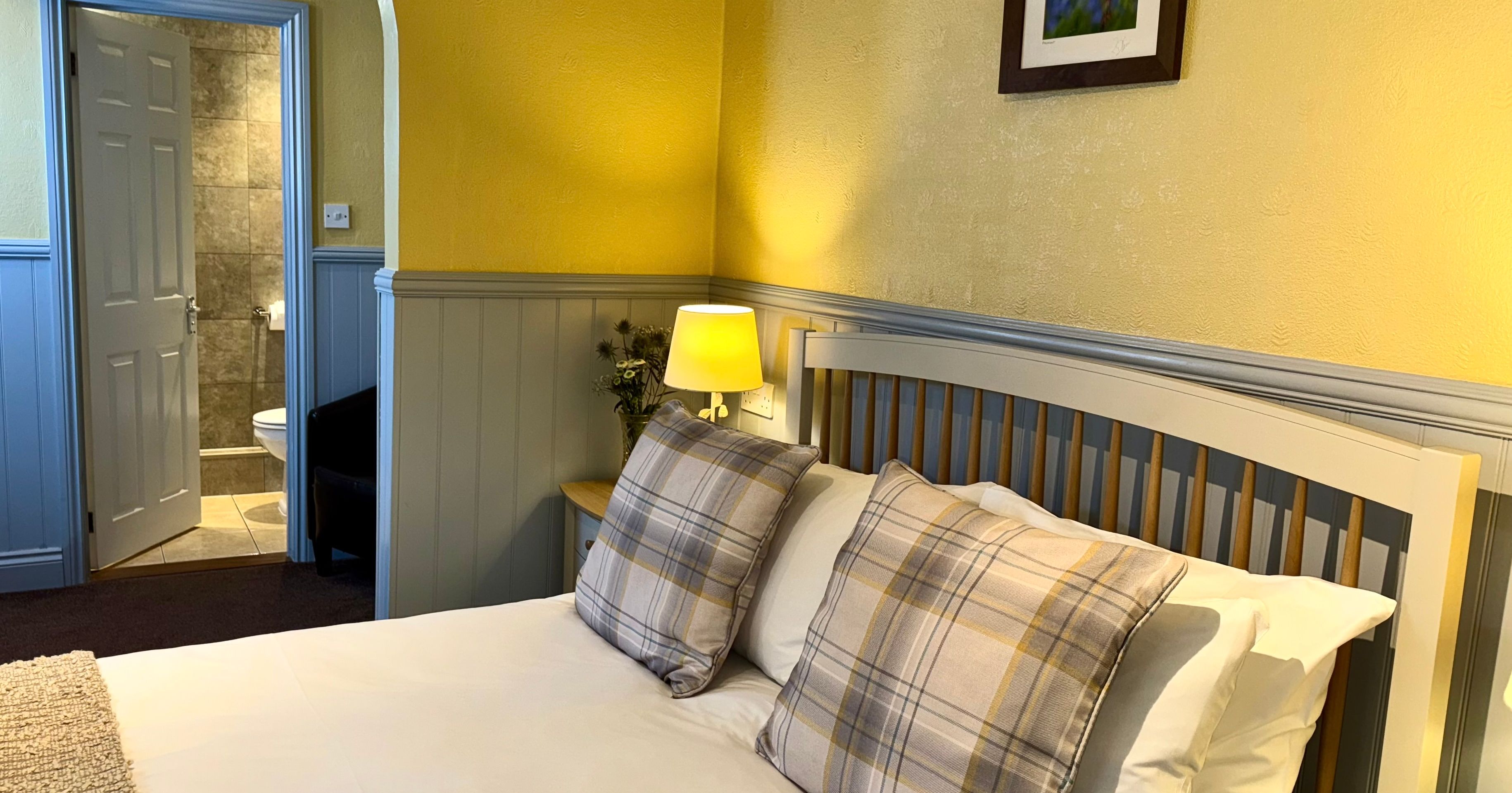 Cozy bedroom with a double bed, plaid pillows, bedside lamp, and open door to a tiled bathroom.